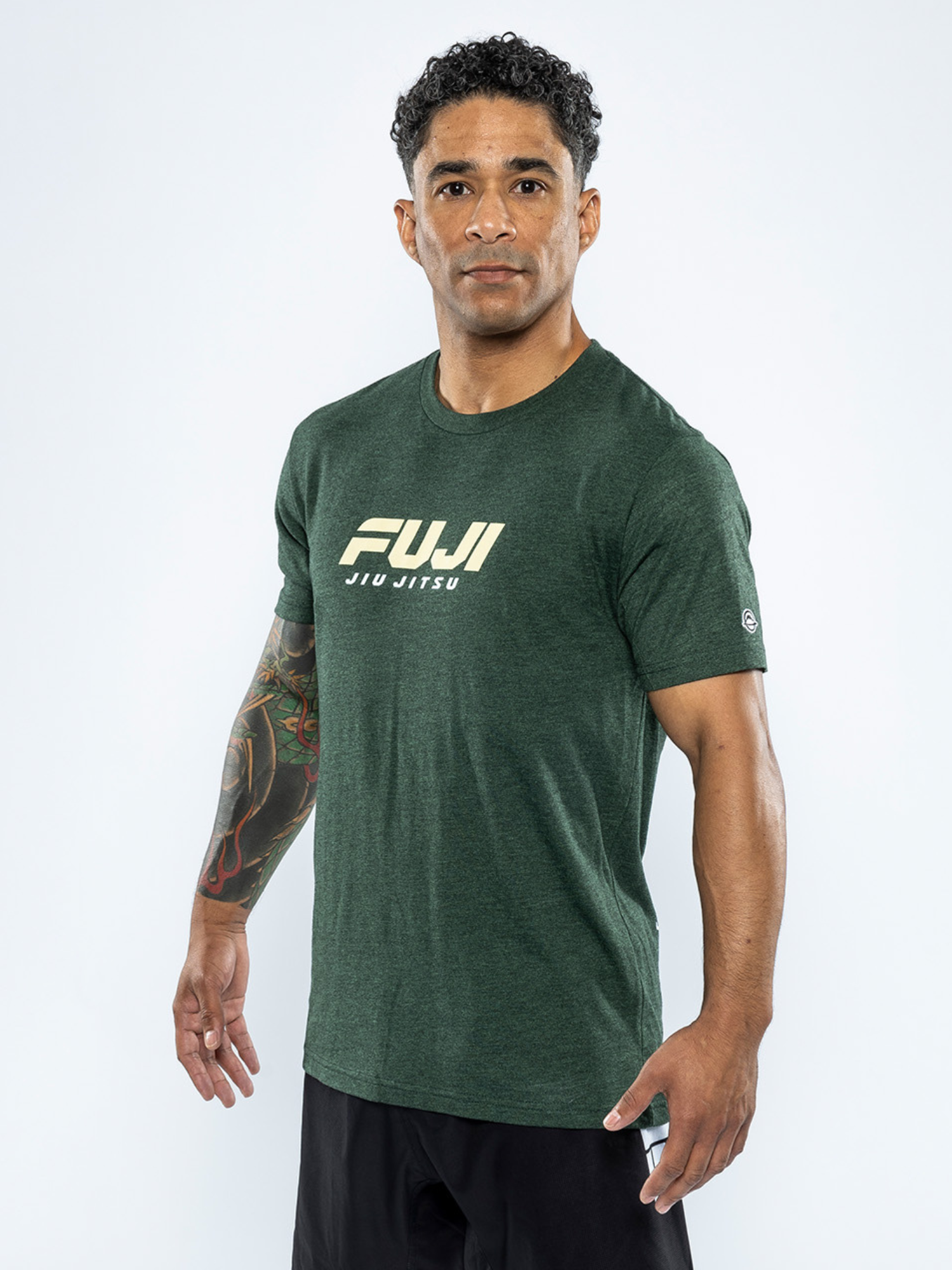 A man with short curly hair and a tattooed right arm wears the Apex T-Shirt Forest Green, featuring "FUJI JIU JITSU" in white lettering, standing against a plain white background.