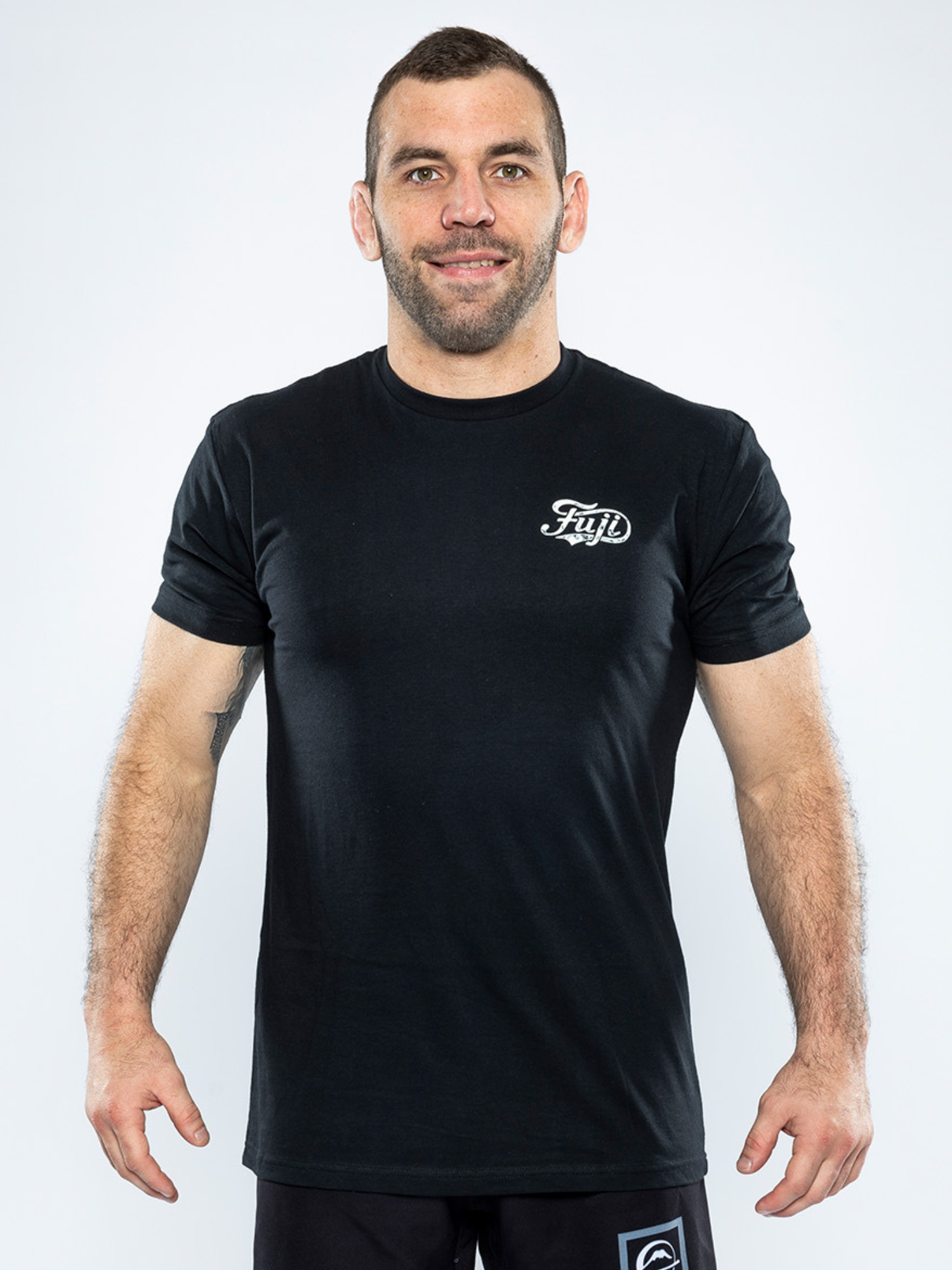 A man stands against a plain white background, smiling and facing forward, wearing the Jiu Jitsu Flow T-Shirt Black with a small white Fuji logo on the upper left chest and black shorts, his arms relaxed at his sides.