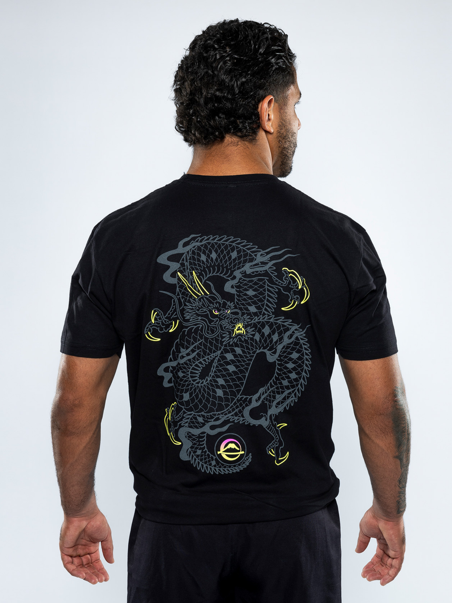 A man with curly hair faces away, wearing the Ryujin T-Shirt Black, which features a large, stylized gray and yellow dragon graphic on the back with fierce details and extended claws.