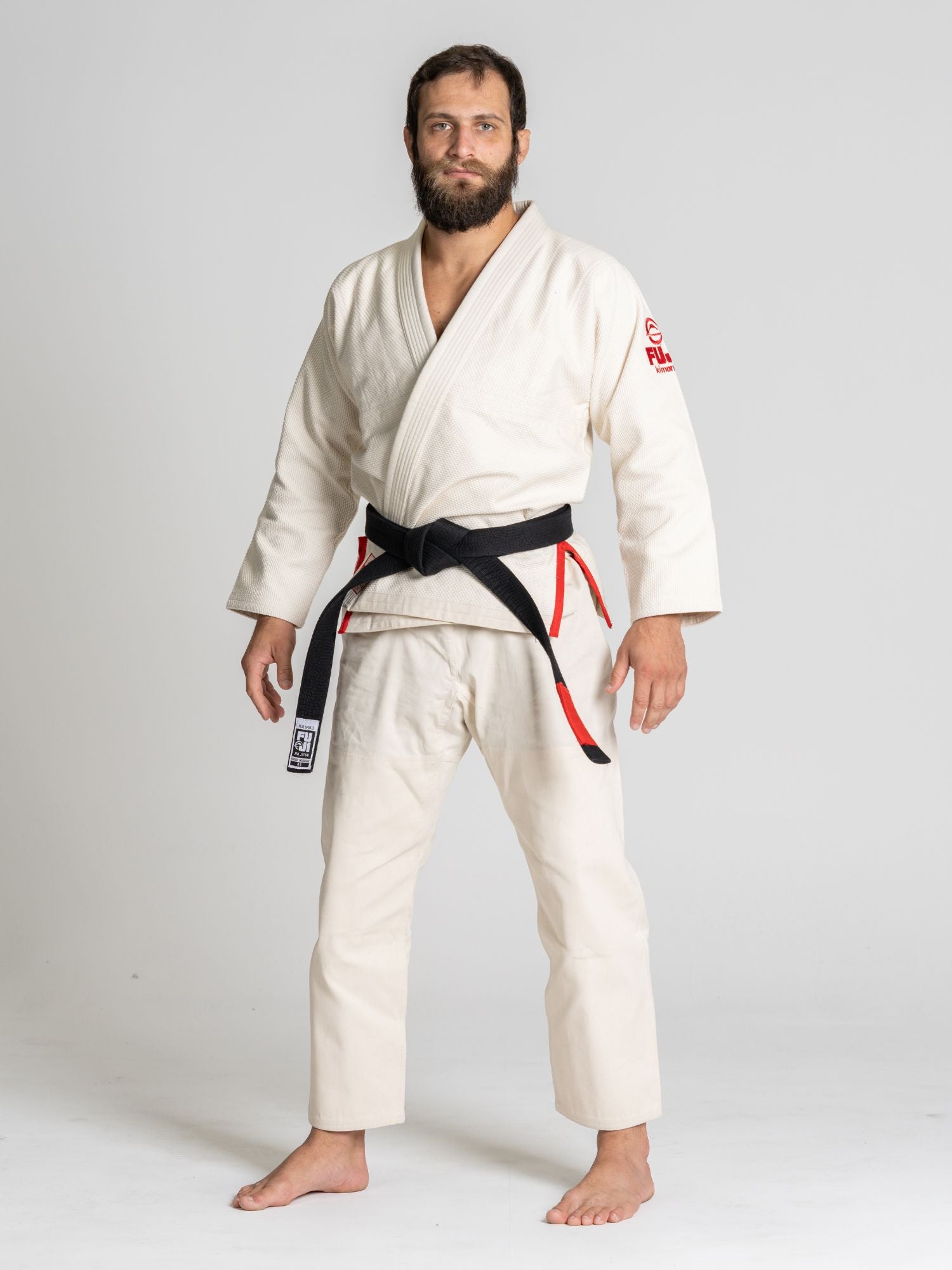A man with a beard stands barefoot on a white floor, wearing the All Around BJJ Gi Natural and a black belt with red stripes, against a plain white background.