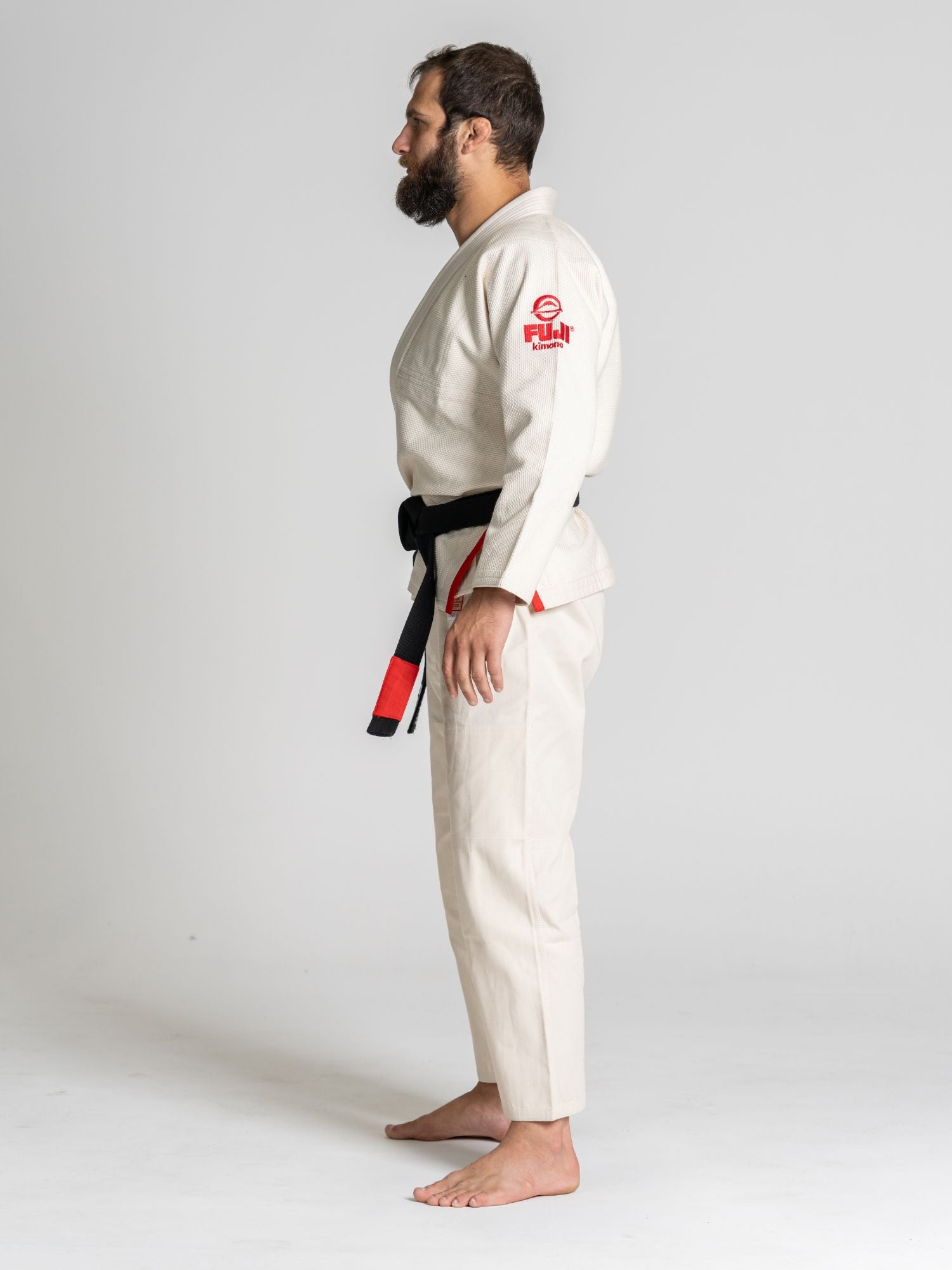 A bearded man stands barefoot in profile, wearing the All Around BJJ Gi Natural—a white gi with a black belt and red FUJI logo on the sleeve. The background is plain and light-colored.