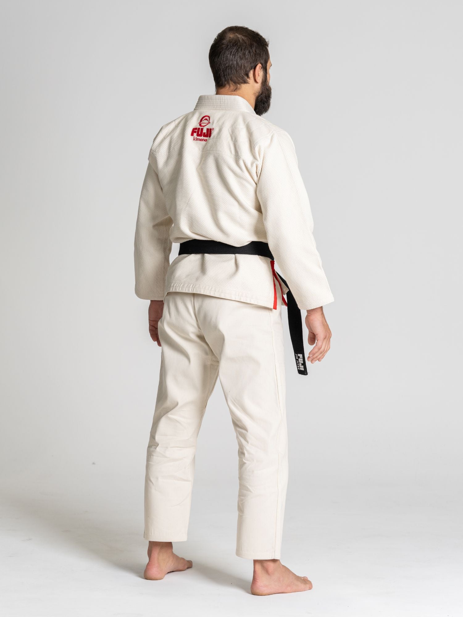 A barefoot, bearded person faces away, wearing the All Around BJJ Gi Natural—an off-white martial arts gi with a black belt, red logo, and "FUJI" on the back—set against a plain light gray background.