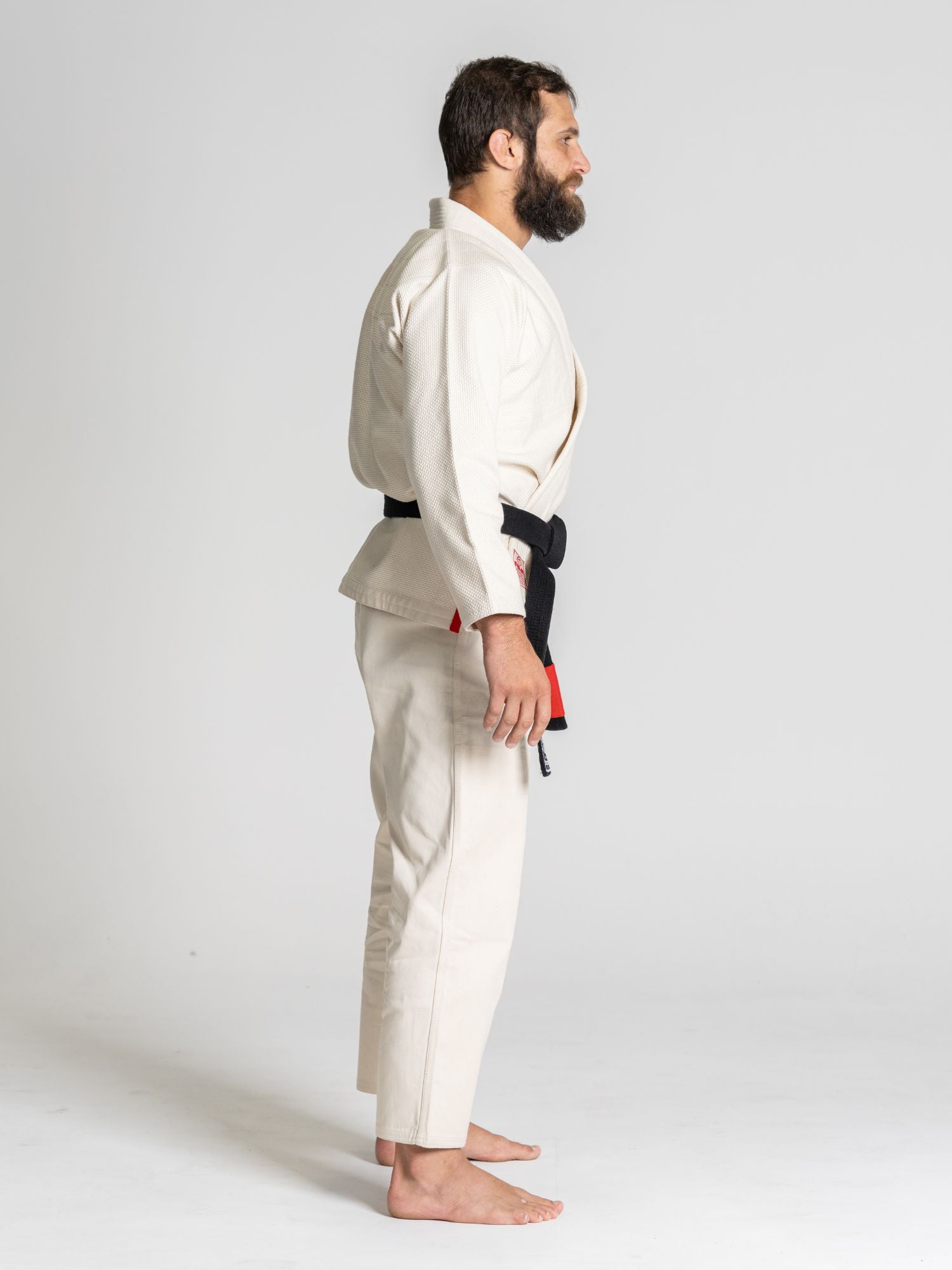 A bearded man stands barefoot in profile, wearing the All Around BJJ Gi Natural and a black belt, against a plain light gray background.
