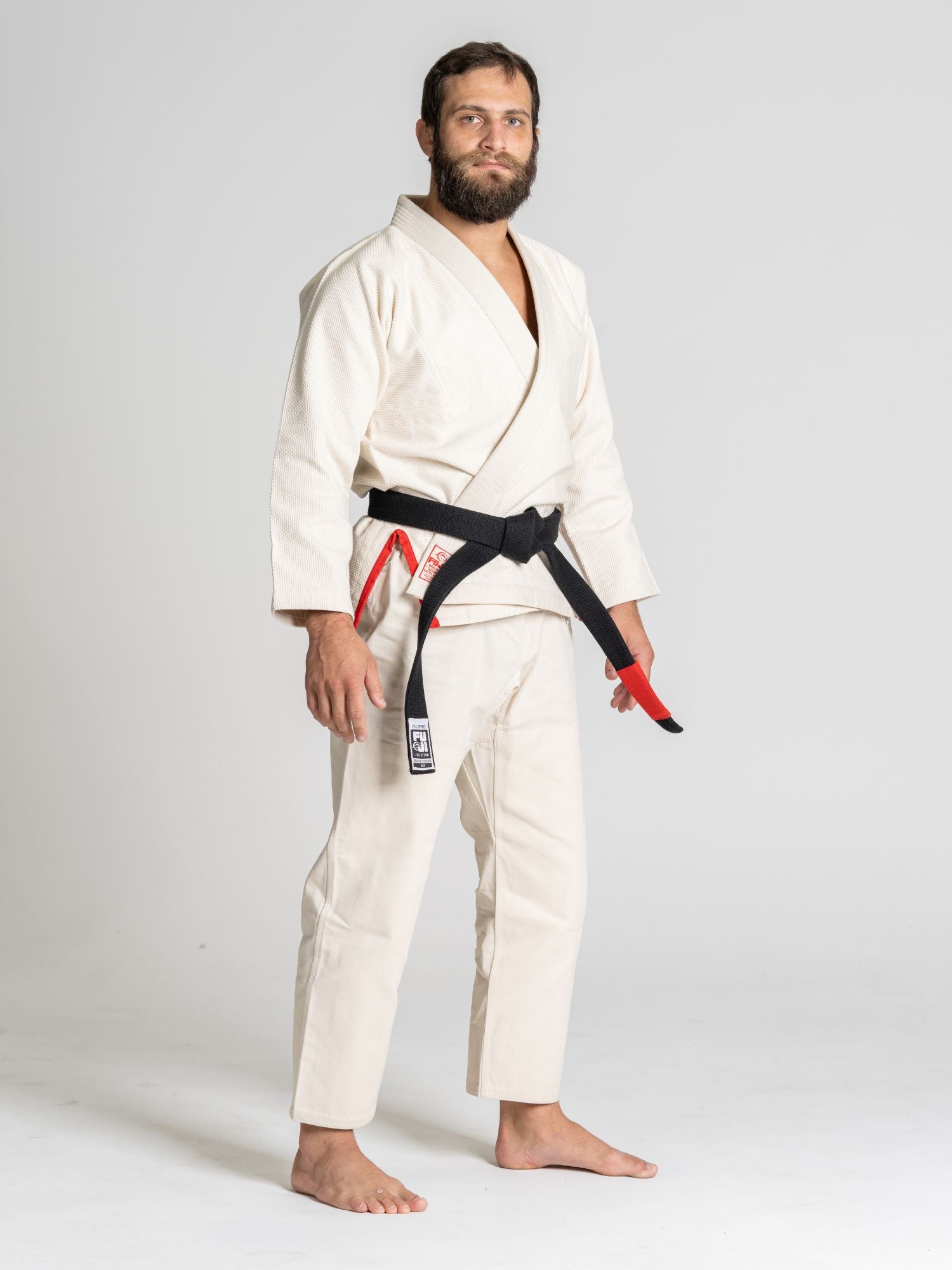 A man with a beard, barefoot and wearing the All Around BJJ Gi Natural with a black belt, stands on a light background and looks at the camera with a neutral expression.
