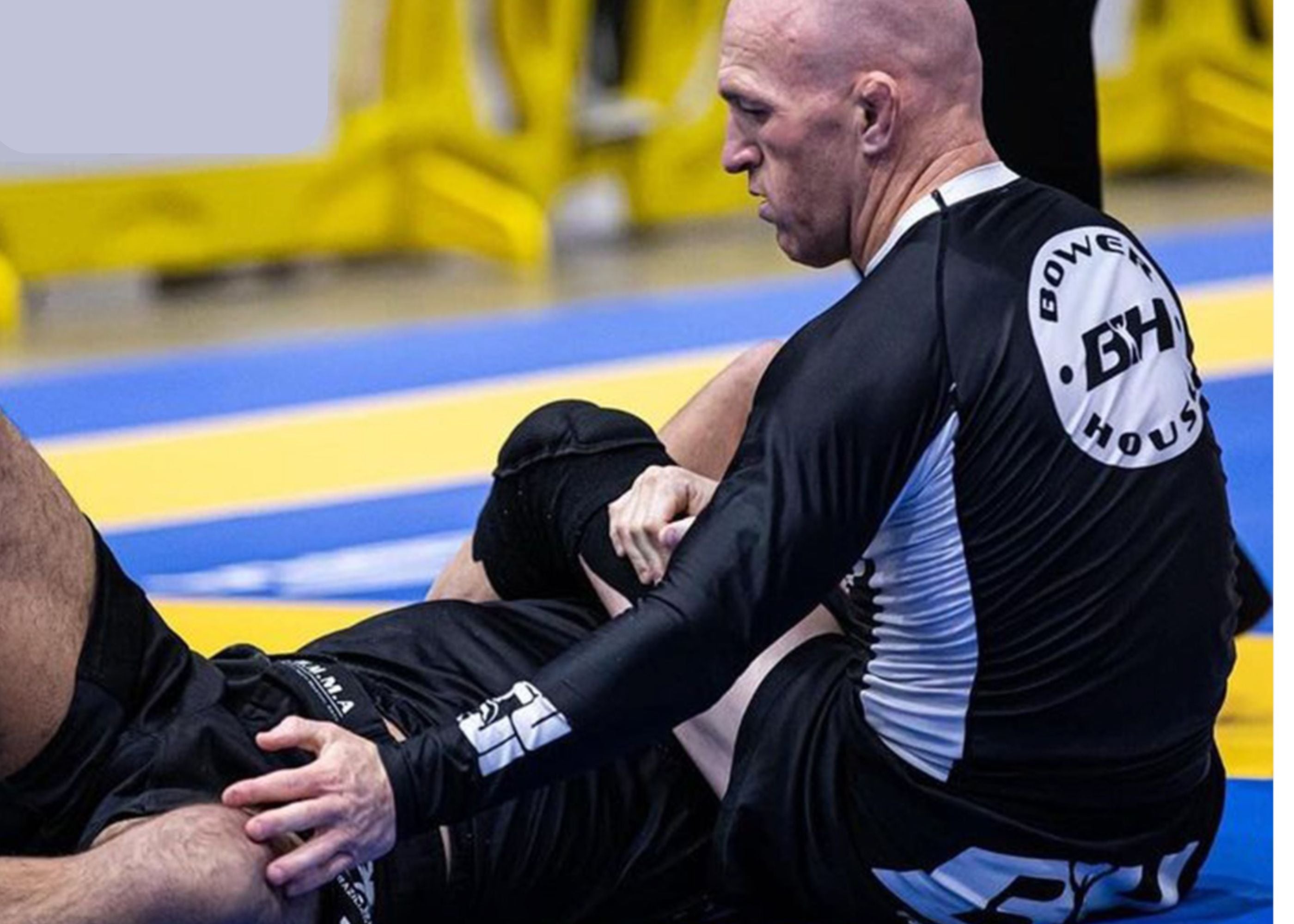 A grappler wearing a black and white rash guard and shorts is applying a submission hold to an opponent during a Brazilian jiu-jitsu match on blue and yellow mats.