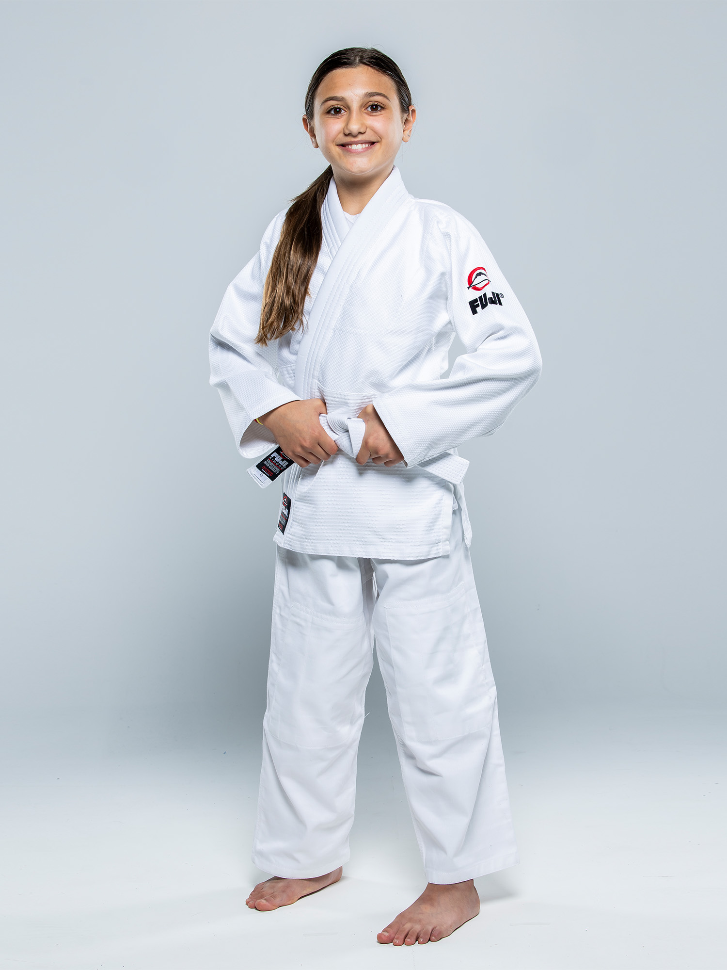 A young girl smiles confidently in a Single Weave Judo Gi White with FUJI branding, standing barefoot against a plain light background and holding her belt.