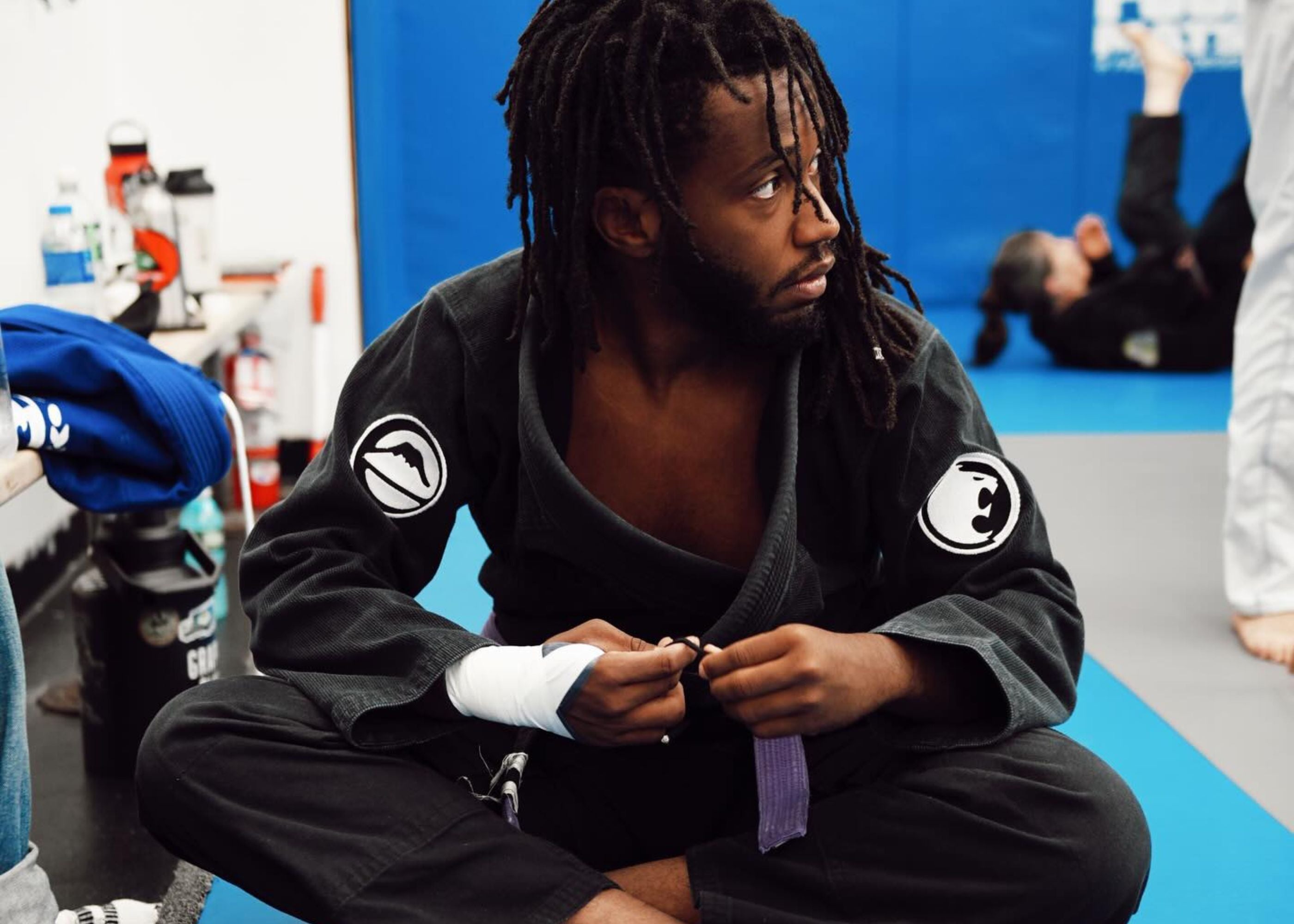 A man in a black martial arts gi sits cross-legged on a mat, wrapping his hand and holding a purple belt. Behind him, other people in gis are training on blue and gray mats.