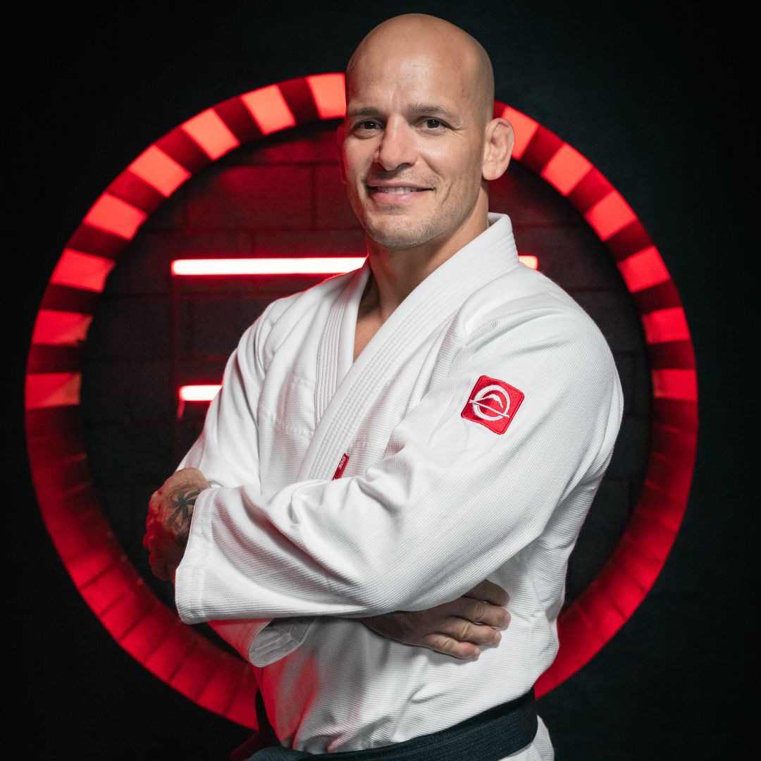A man in a white martial arts gi stands with arms crossed, smiling, in front of a glowing red circular light on a dark background.