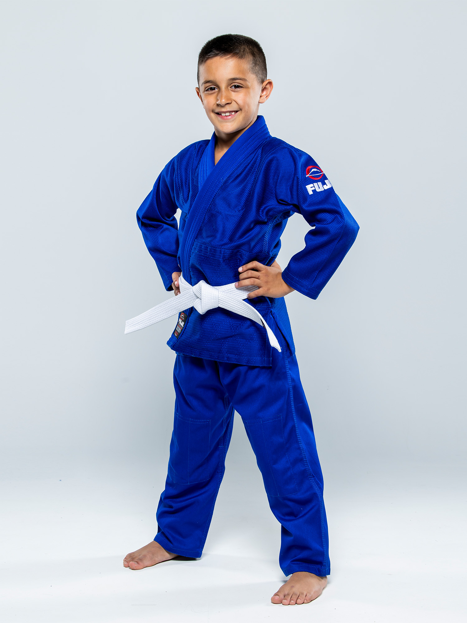 A young boy confidently stands barefoot with his hands on his hips, smiling, wearing the Single Weave Judo Gi Blue and a white belt against a plain light background.