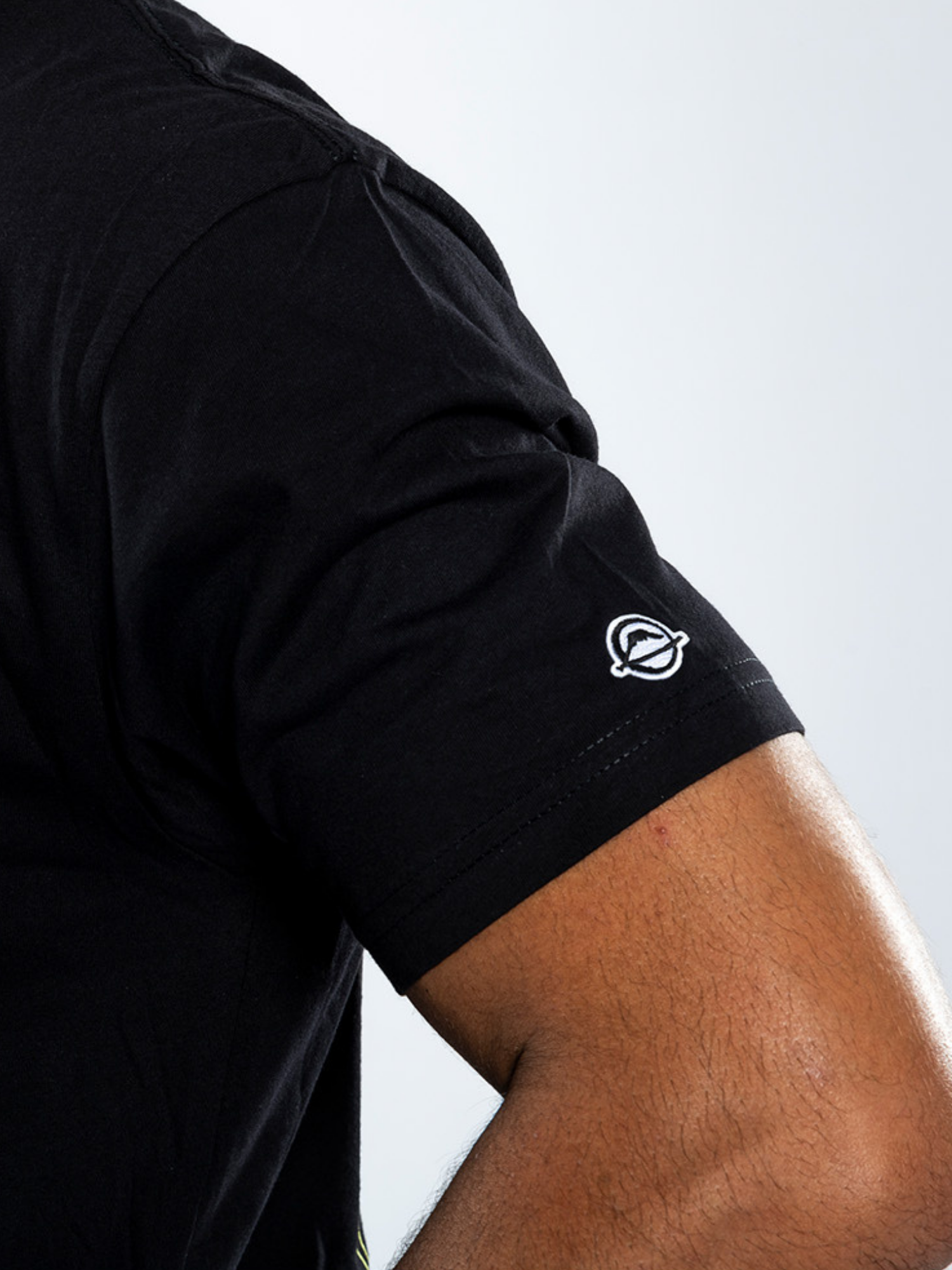 A close-up of a person’s upper arm wearing the Ryujin T-Shirt Black, featuring a small white embroidered planet and ring logo near the sleeve hem.