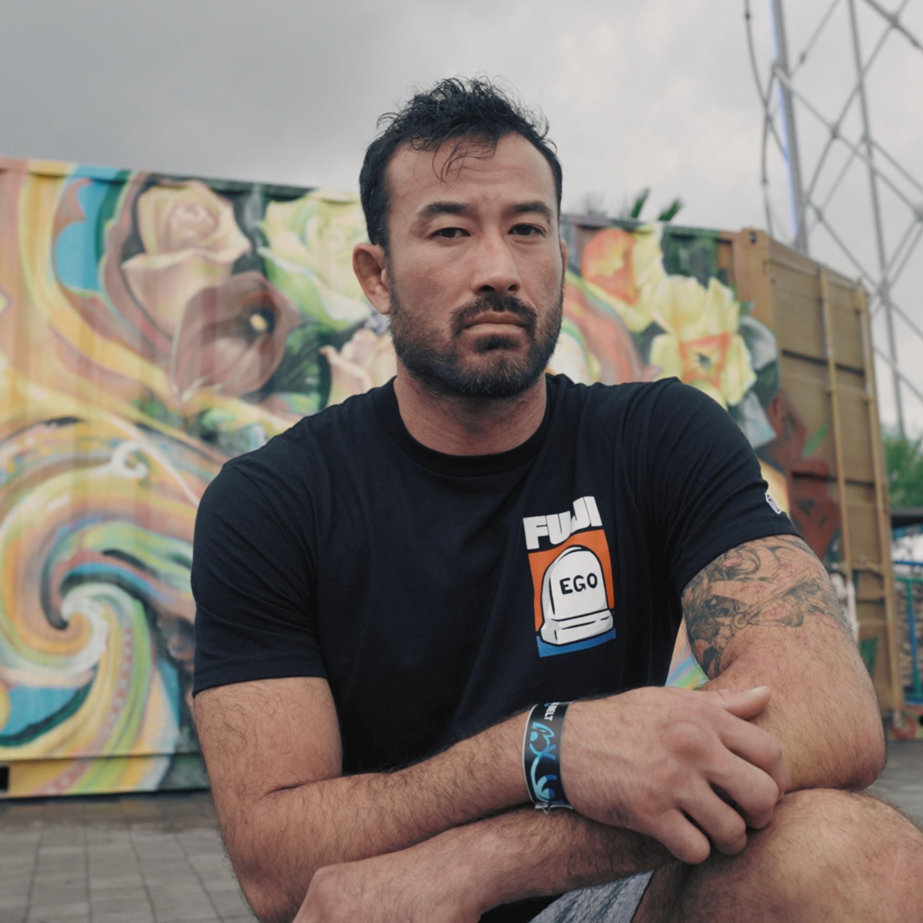 A man with short dark hair and a beard sits outdoors in front of a colorful mural. He wears a black FUJI T-shirt and a wristband, showing tattoos on his left arm, and looks seriously at the camera.