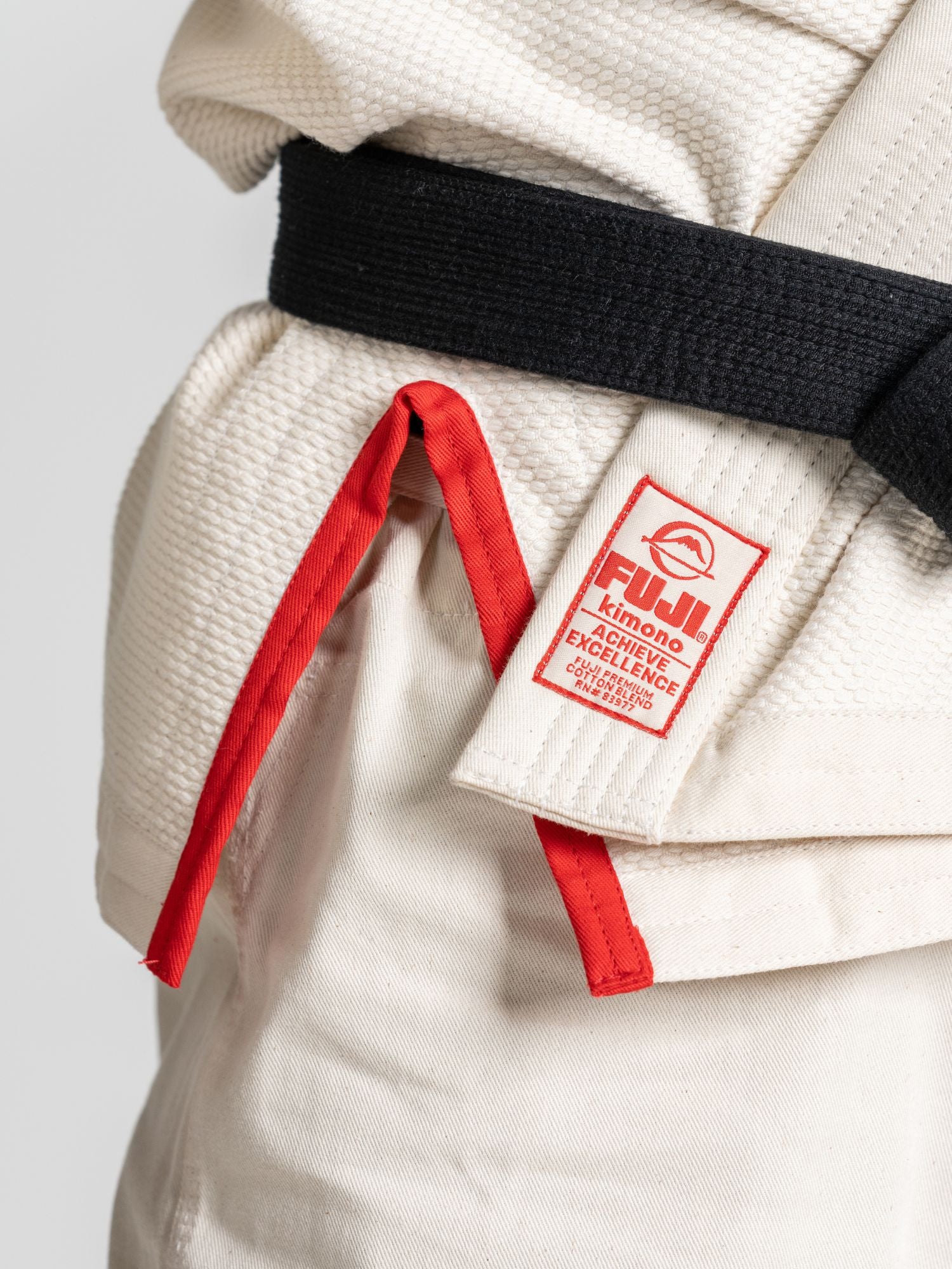 Close-up of the All Around BJJ Gi Natural with a black belt tied at the waist. A red tag with "FUJI Kimono Excellence" is sewn onto the gi, with a red string hanging next to it.