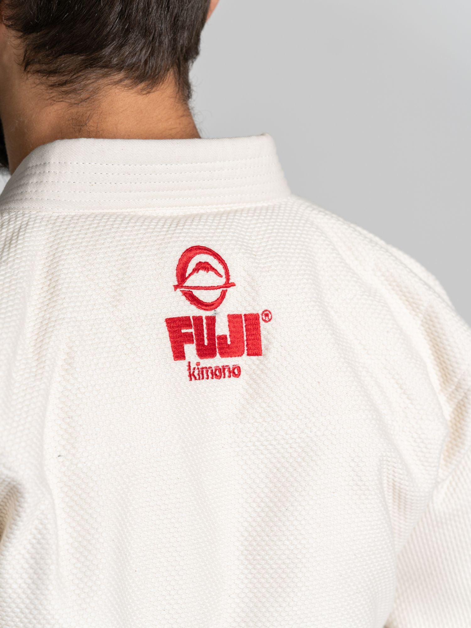 Close-up of a person wearing the All Around BJJ Gi Natural with the red FUJI kimono logo embroidered on the collar. The person is facing away, showing only part of their head and shoulder.
