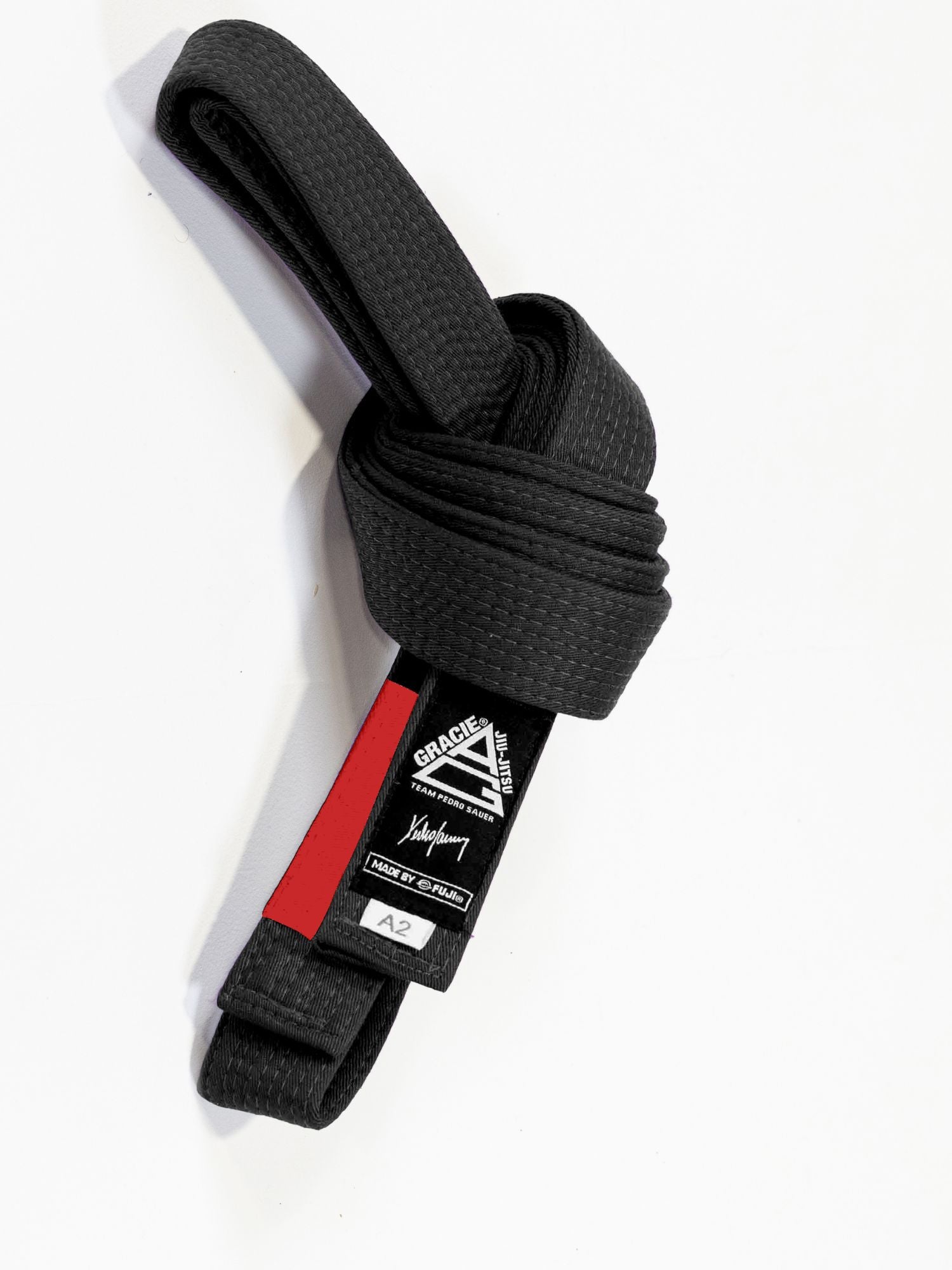 The Pedro Sauer Jiu Jitsu Belt Black, featuring a red and black label and white A2 size tag, is neatly rolled on a white background.