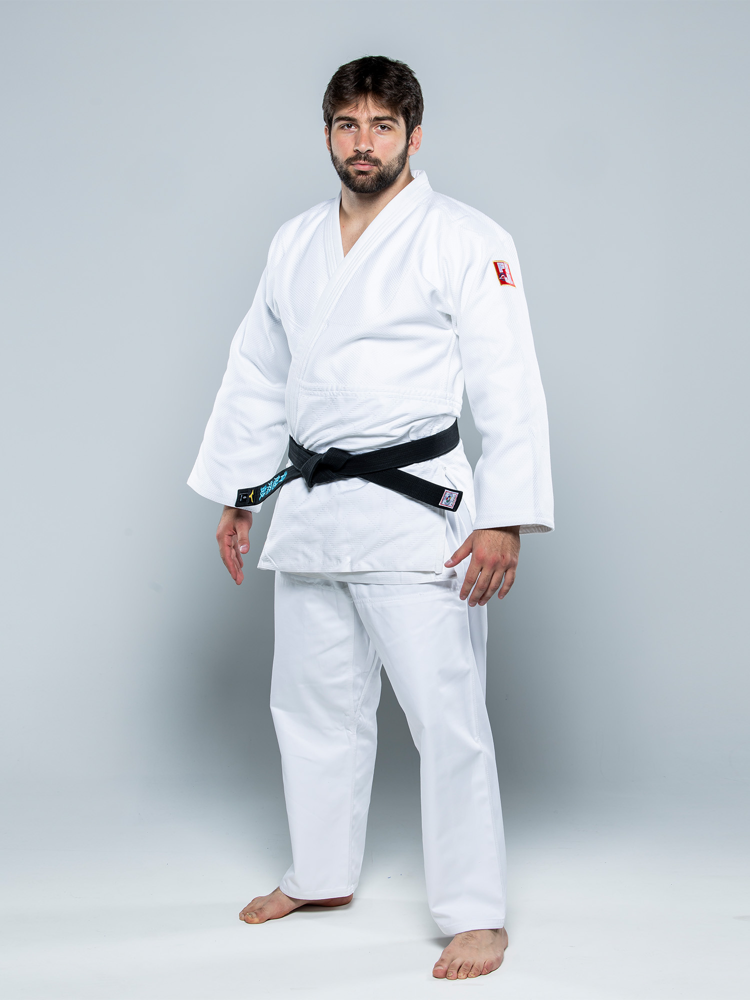A man wears the Euro Competition Judo Gi White with a black belt, standing barefoot on a light gray background and looking at the camera with a neutral expression.