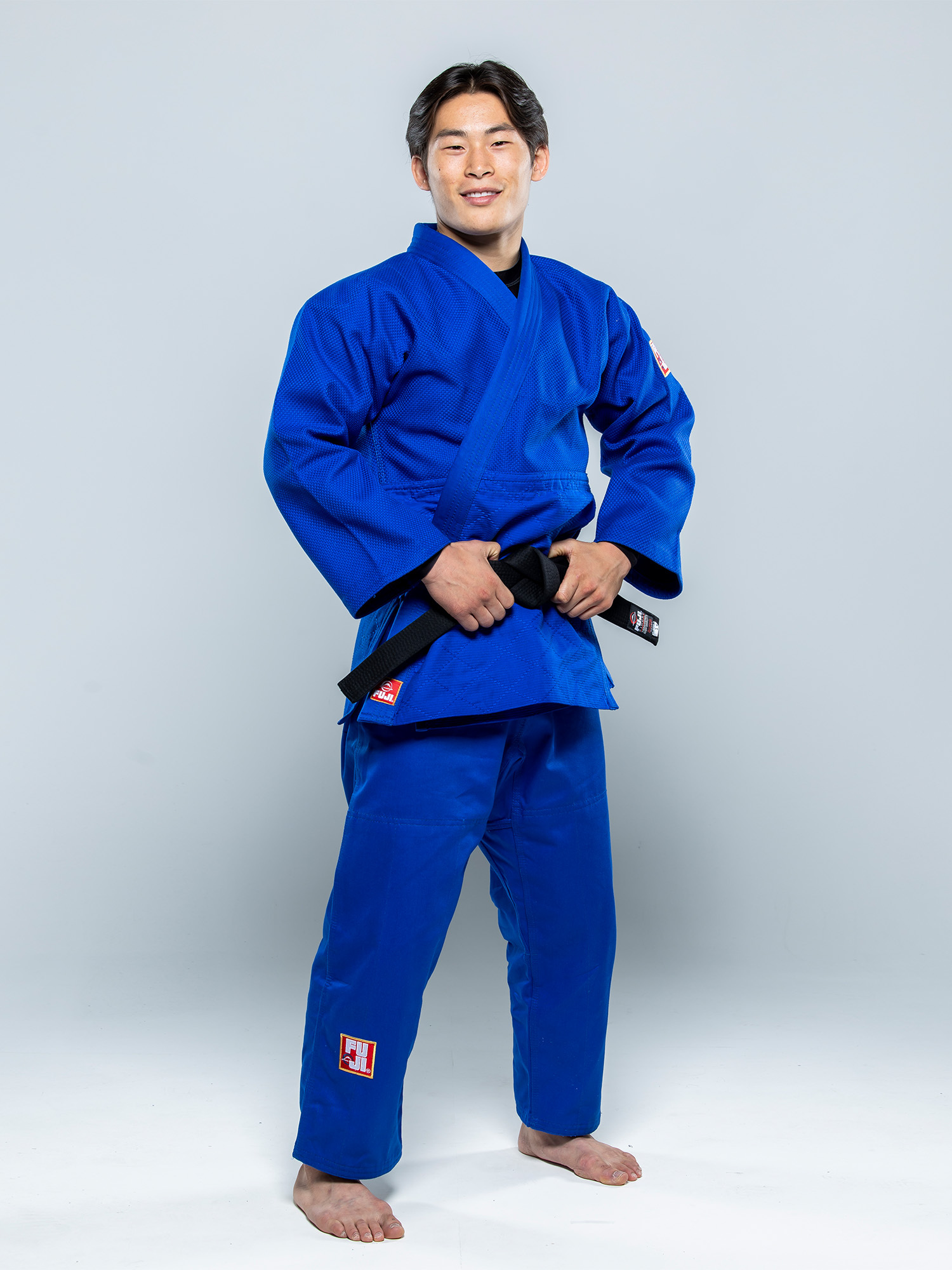 A man stands barefoot, smiling confidently and holding a black belt while wearing the Euro Competition Judo Gi Blue in front of a plain light gray background.