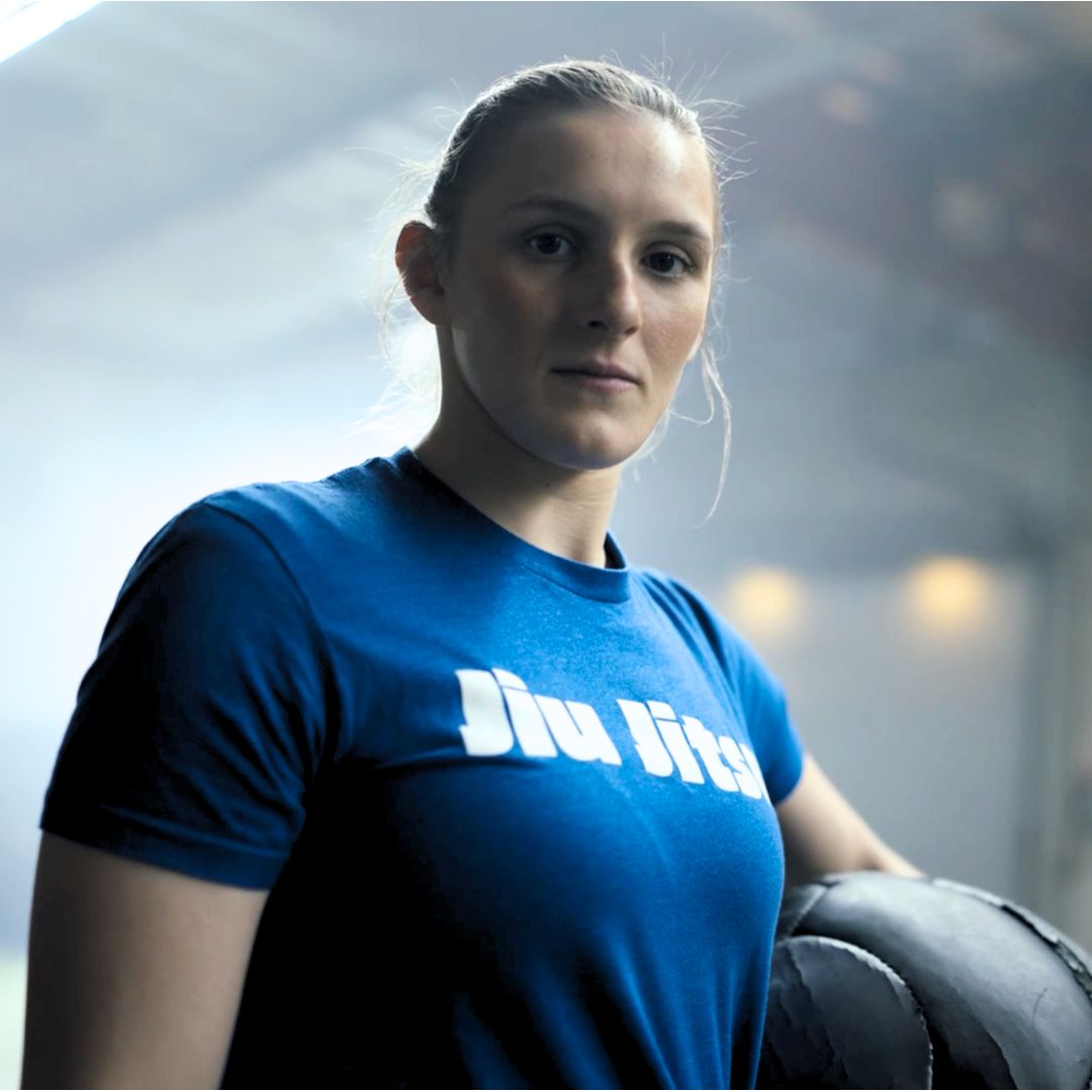 A woman in a blue Jiu Jitsu shirt stands indoors, looking confidently at the camera with one arm resting on black training equipment. The background is softly lit and slightly blurred.