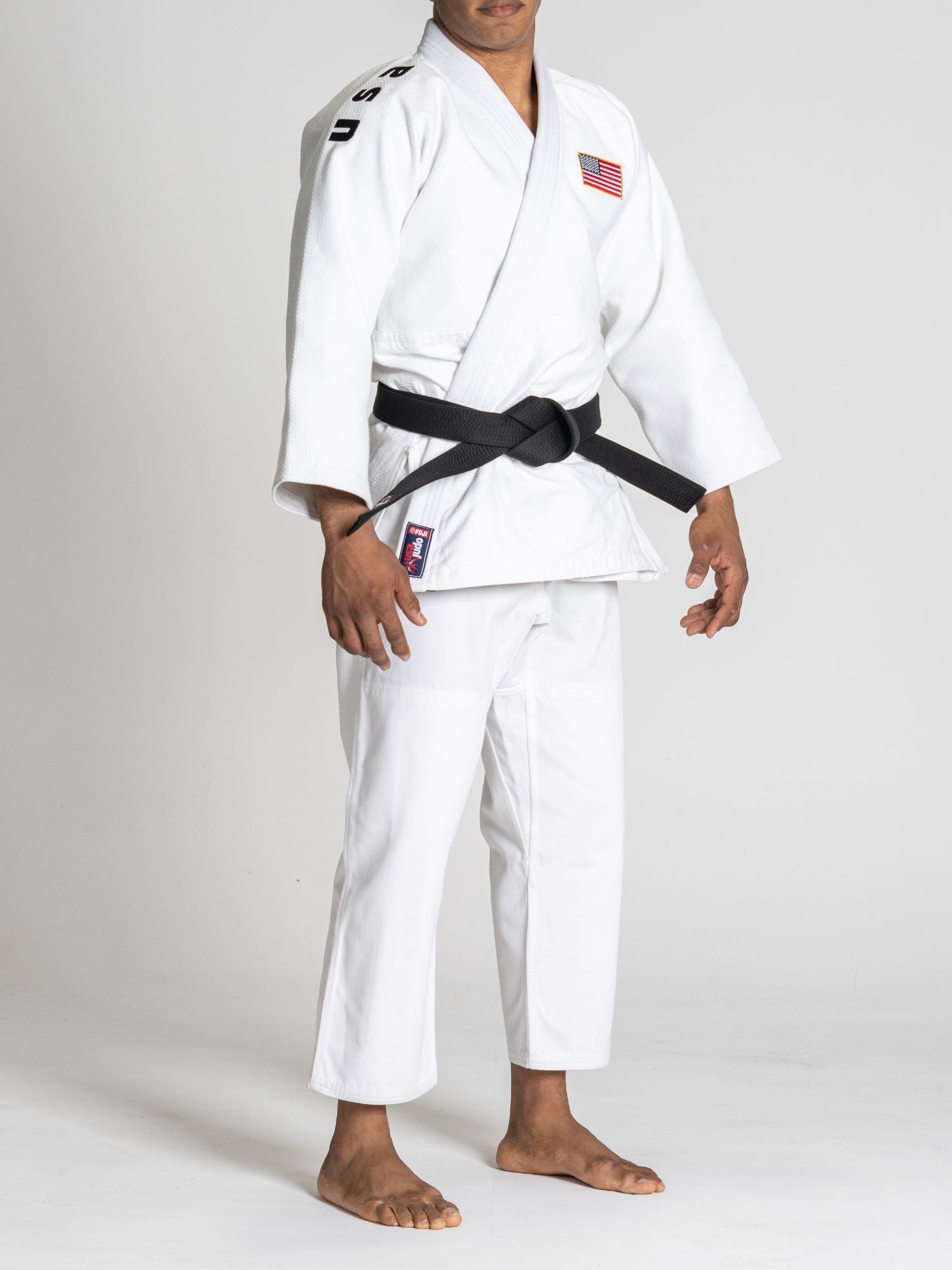 A person stands barefoot against a plain white background, wearing the USA Judo Euro Competition Judo Gi White with a black belt and a USA flag patch on the chest.