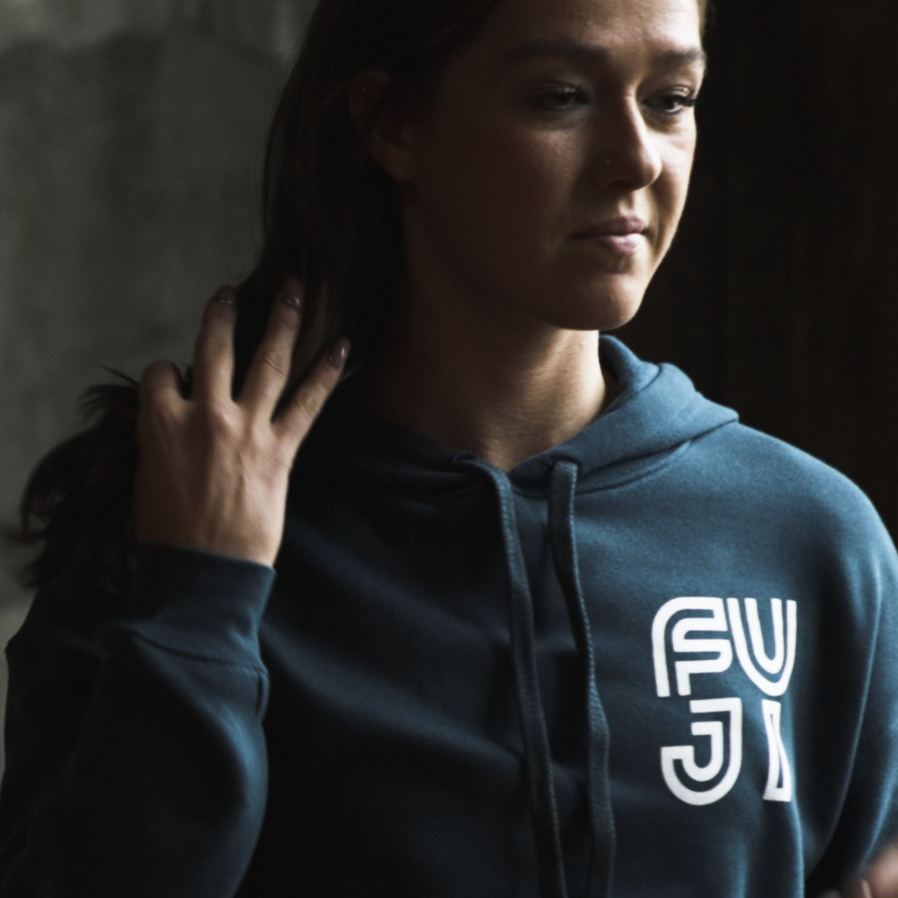 A person wearing a dark hoodie with the white text FUJI on it stands indoors, touching their hair and looking slightly downward. The background is dimly lit and out of focus.