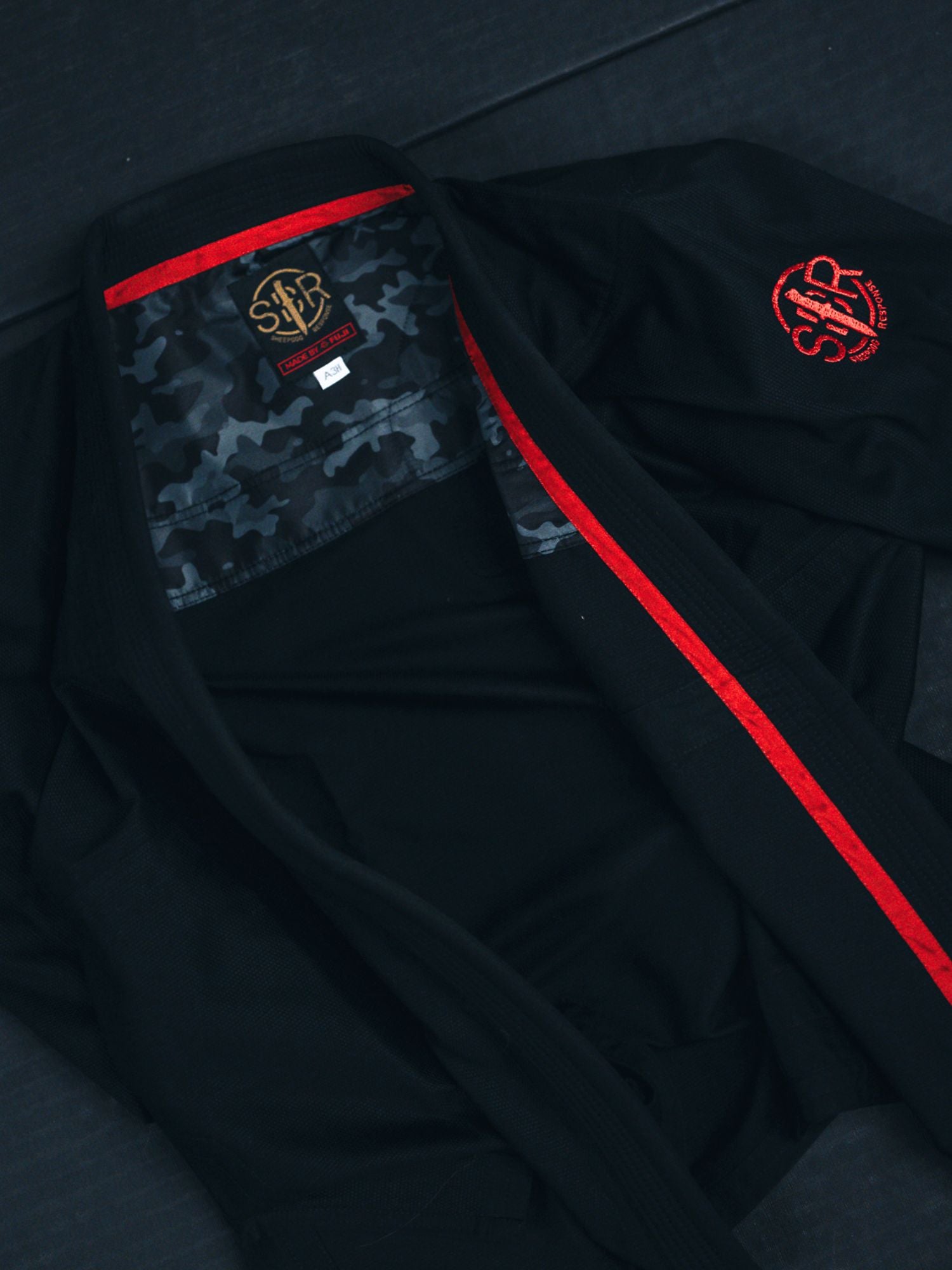 The Sheepdog Response Standard Suparaito BJJ Gi Black features red trim and embroidery, and is laid open to reveal its camo-patterned inner lining and a visible label on the collar.