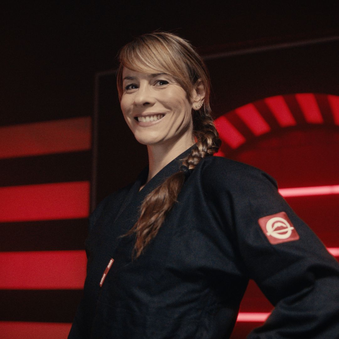 A woman with light brown hair in a braid smiles confidently while wearing a dark martial arts uniform with a red patch on the sleeve, standing in front of a background with red light accents.