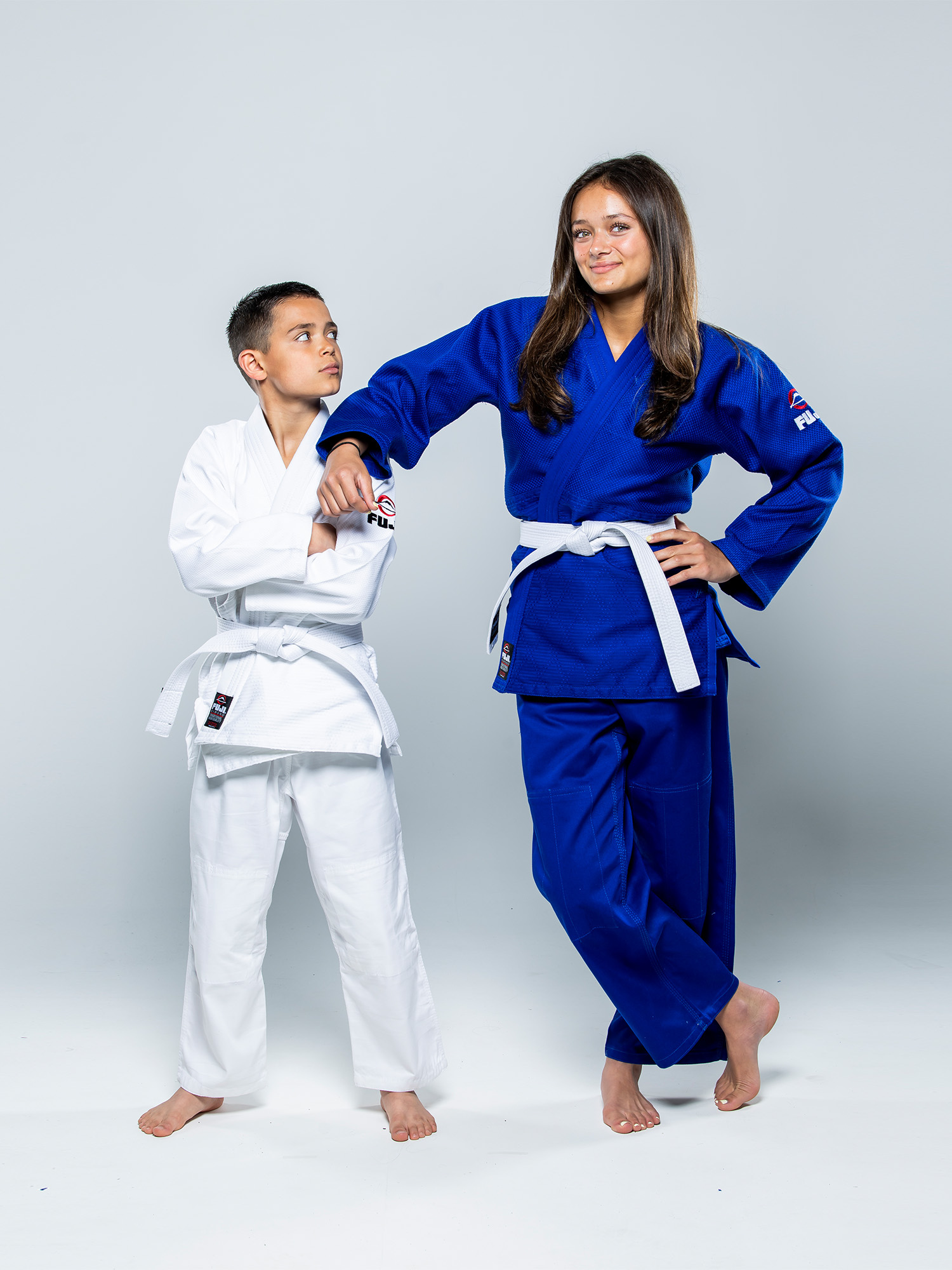 Two young martial artists pose confidently. The girl wears the Single Weave Judo Gi Blue with a white belt and stands with her hand on her hip, resting her arm on the shoulder of a boy in a white gi. Both are barefoot against a plain background.