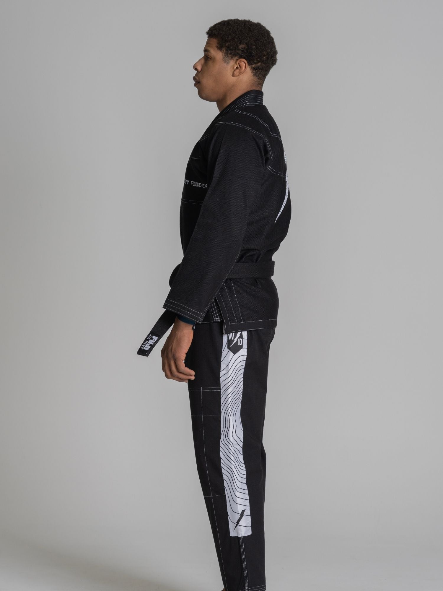 A man stands in profile wearing the We Defy Topo Suparaito BJJ Gi Black, featuring white graphic designs on the pants and a black belt, against a plain gray background.