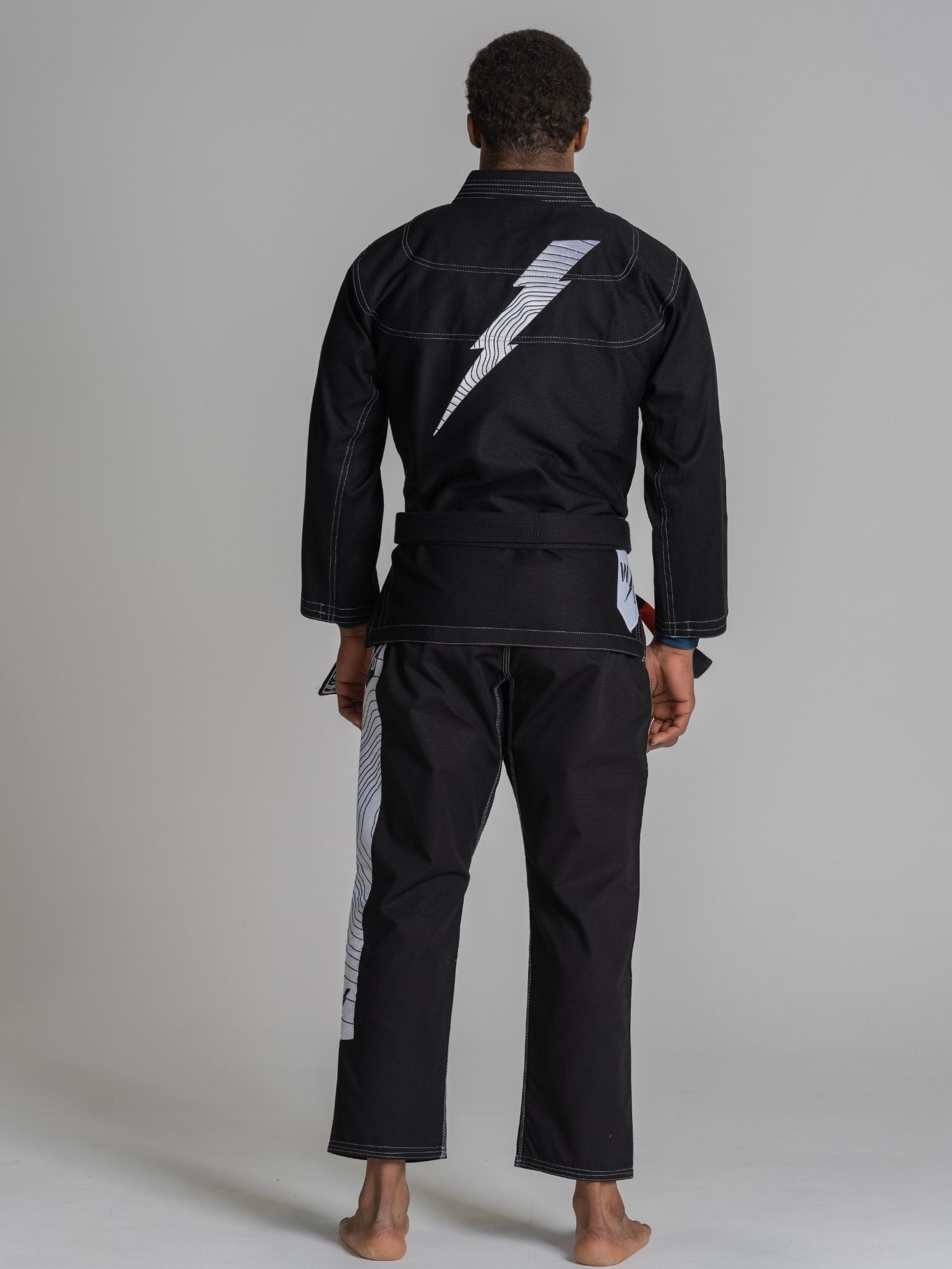 A person stands barefoot facing away, wearing the We Defy Topo Suparaito BJJ Gi Black featuring a large white lightning bolt on the back and a matching side stripe, set against a simple gray background.