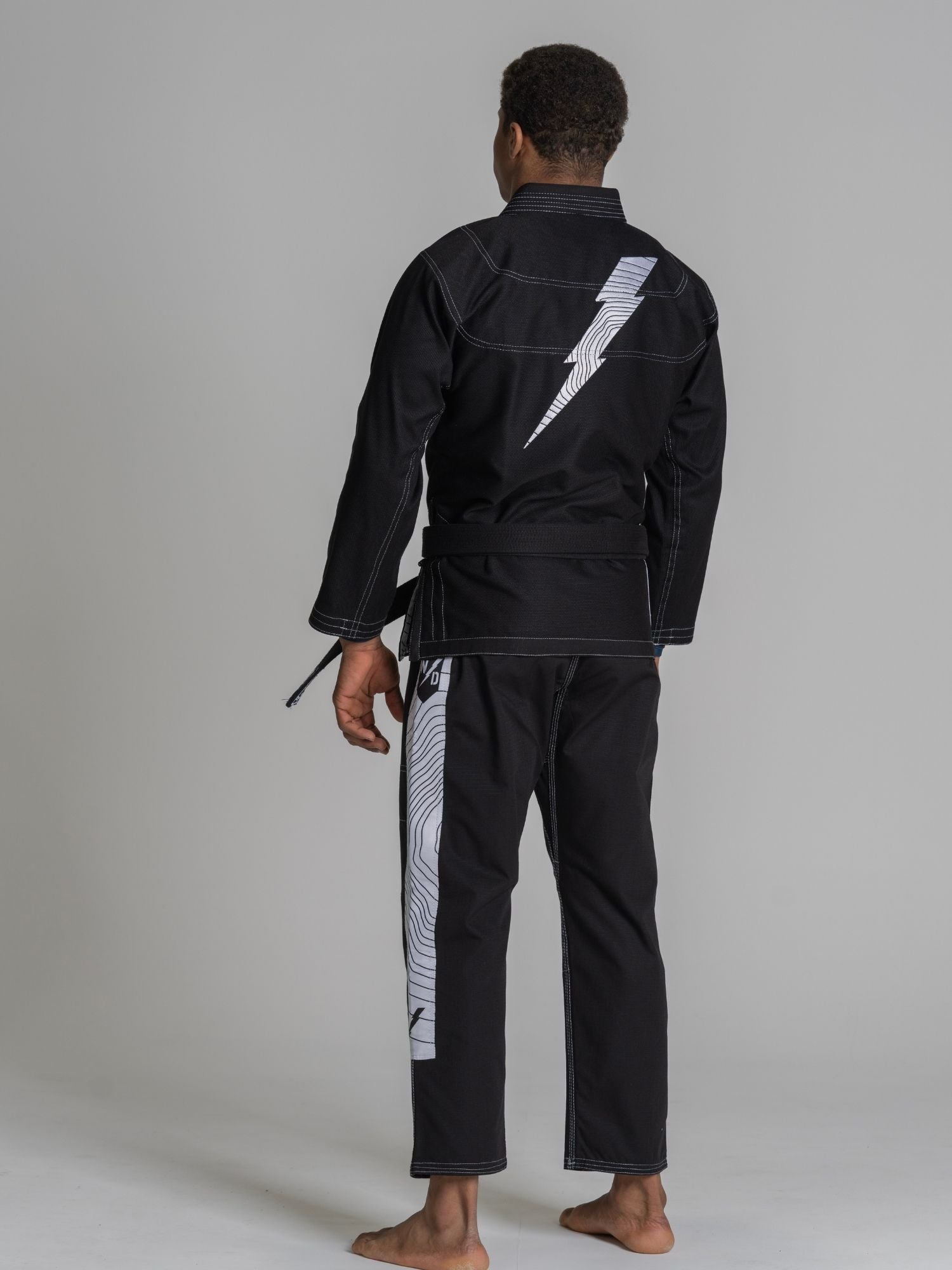 A person stands barefoot facing away, wearing the We Defy Topo Suparaito BJJ Gi Black with a white lightning bolt design on the back and white patterned stripes on the pants, set against a plain gray background.