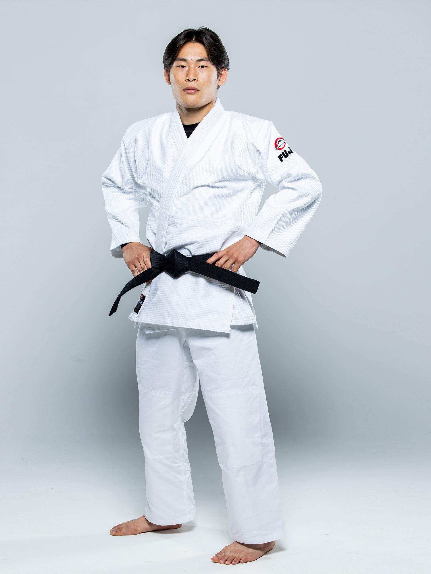 A man models the Single Weave Judo Gi White with a black belt, standing barefoot on a white floor, hands on hips and looking confidently at the camera.
