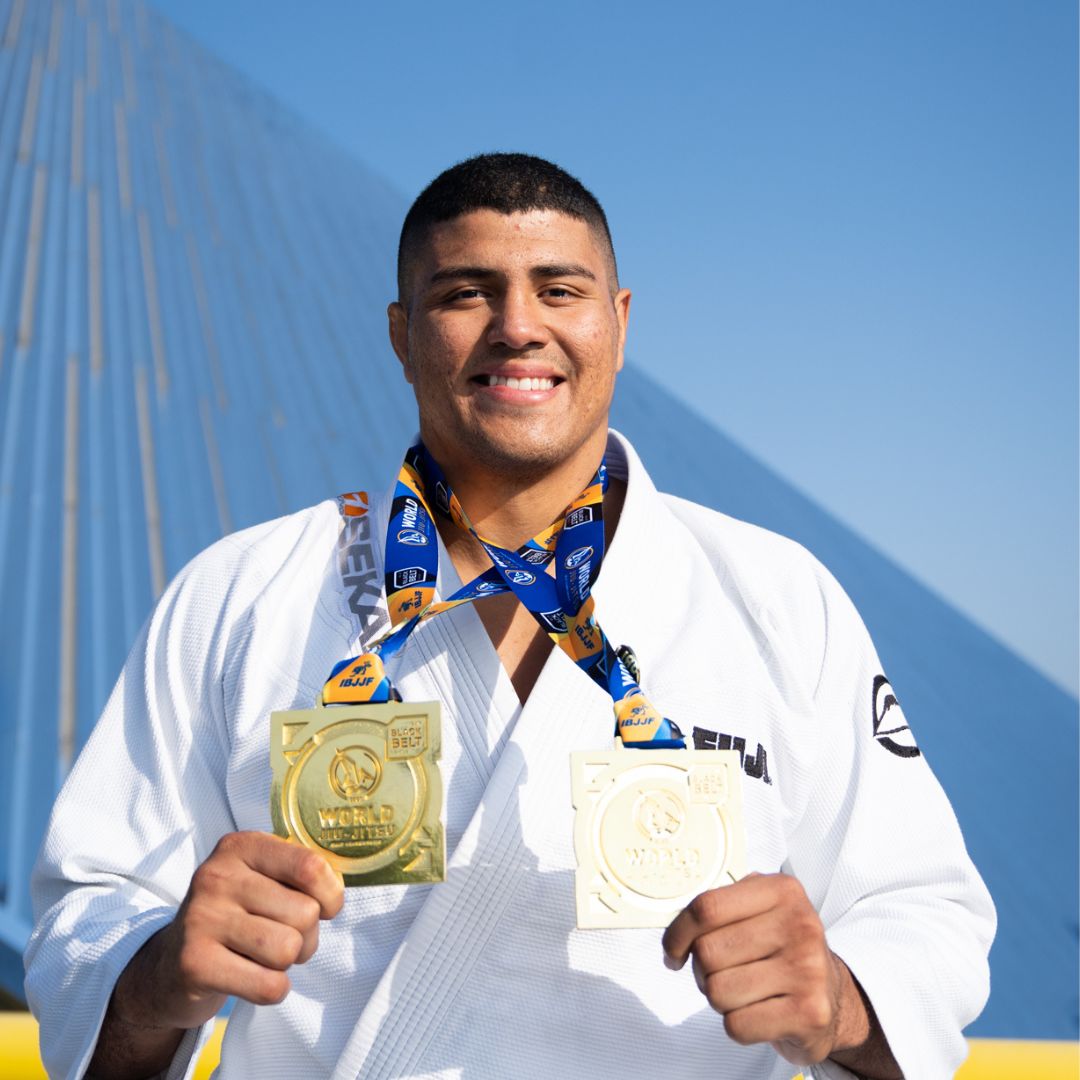 A smiling male judo athlete in a white gi holds two gold medals, wearing a medal ribbon around his neck, with a modern bridge structure and blue sky in the background.