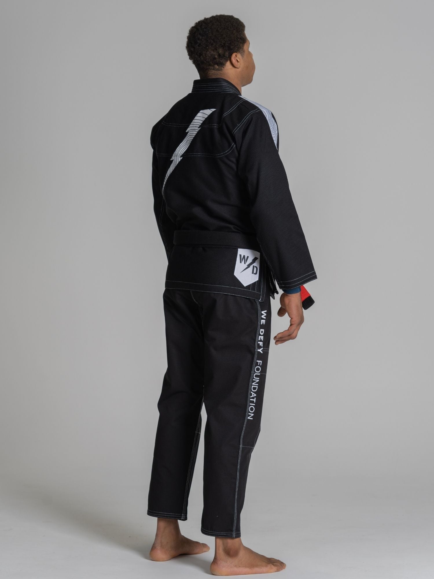 A person stands barefoot on a plain background, wearing the We Defy Topo Suparaito BJJ Gi Black with white stitching, a black belt, white stripe design on the back, and "WOLDRY FOUNDATION" along the side of the pants.