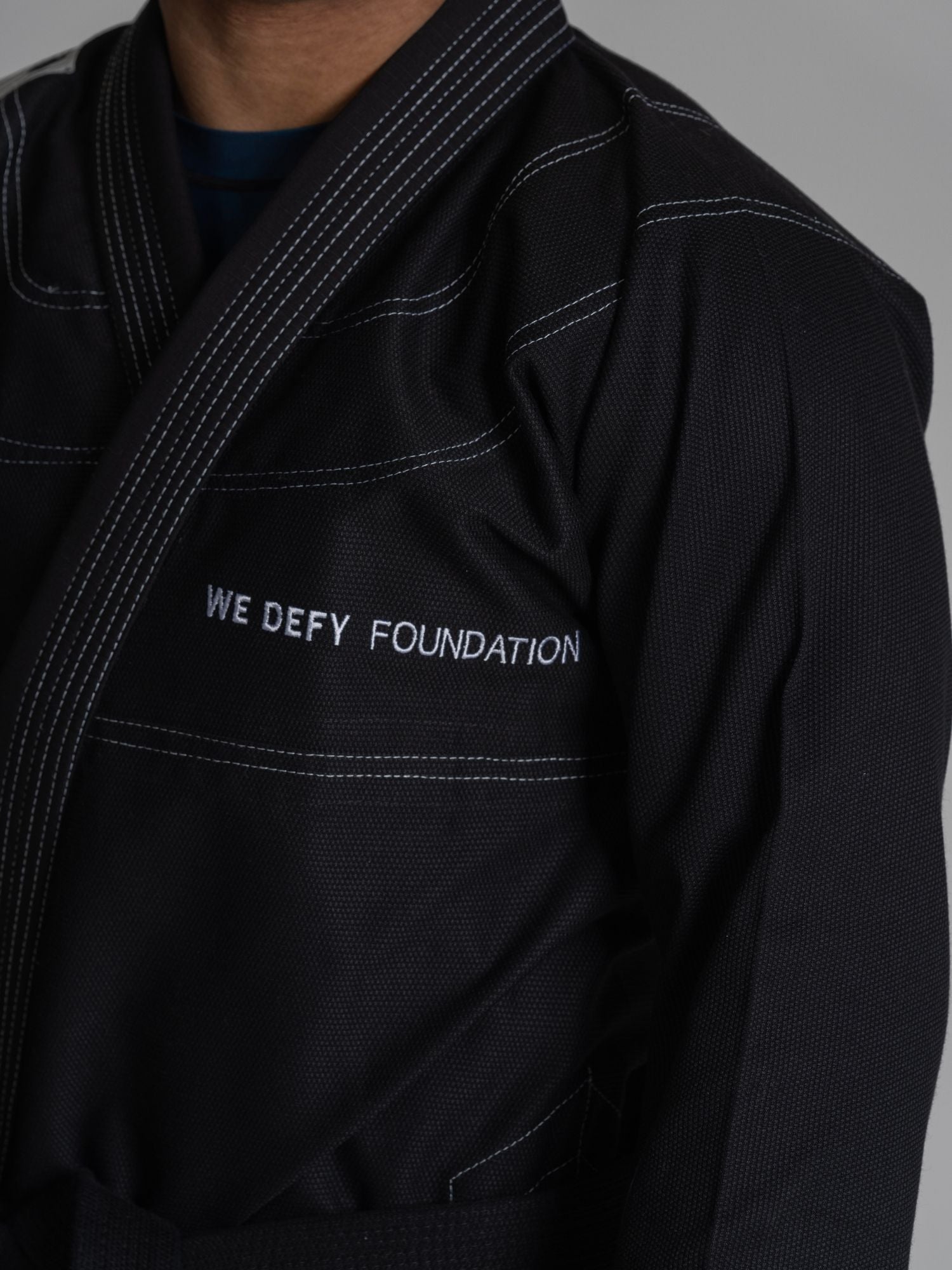 A person wears the We Defy Topo Suparaito BJJ Gi Black, featuring white stitching and "WE DEFY FOUNDATION" embroidered in white on the chest.