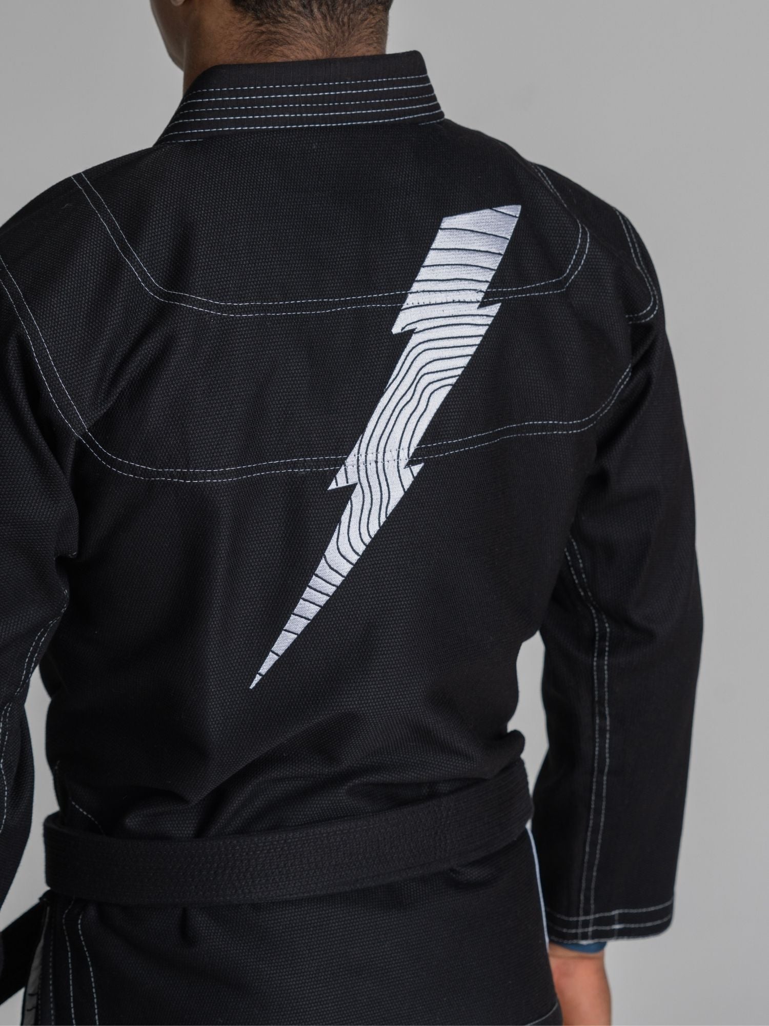 A person is wearing the We Defy Topo Suparaito BJJ Gi Black, which features white stitching and a large white lightning bolt design on the back. The image shows their back, part of their arm, and waist.