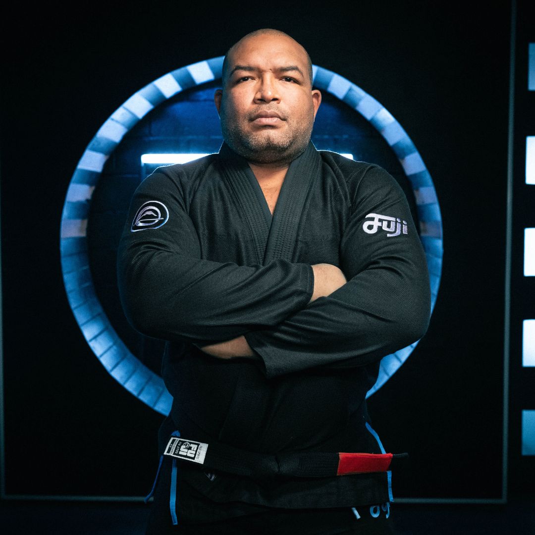 A man wearing a black Brazilian Jiu-Jitsu gi stands with arms crossed in front of a glowing circular light, looking confidently at the camera.