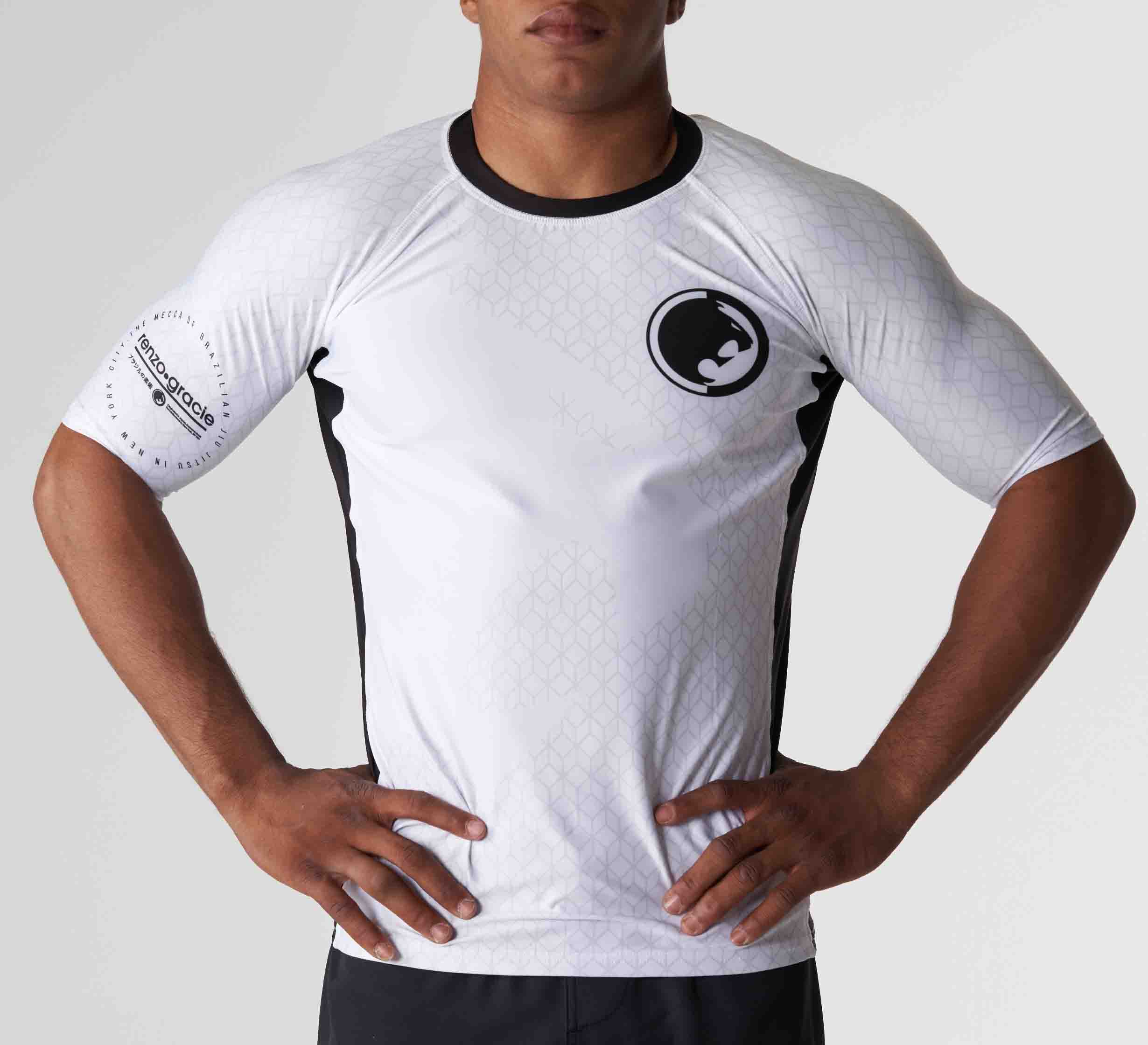 A person stands with hands on hips, wearing the Renzo Gracie Mecca Ranked Short Sleeve Rashguard—a fitted black and white athletic shirt with a circular logo on the chest and sponsor logo on the sleeve—against a plain light background.