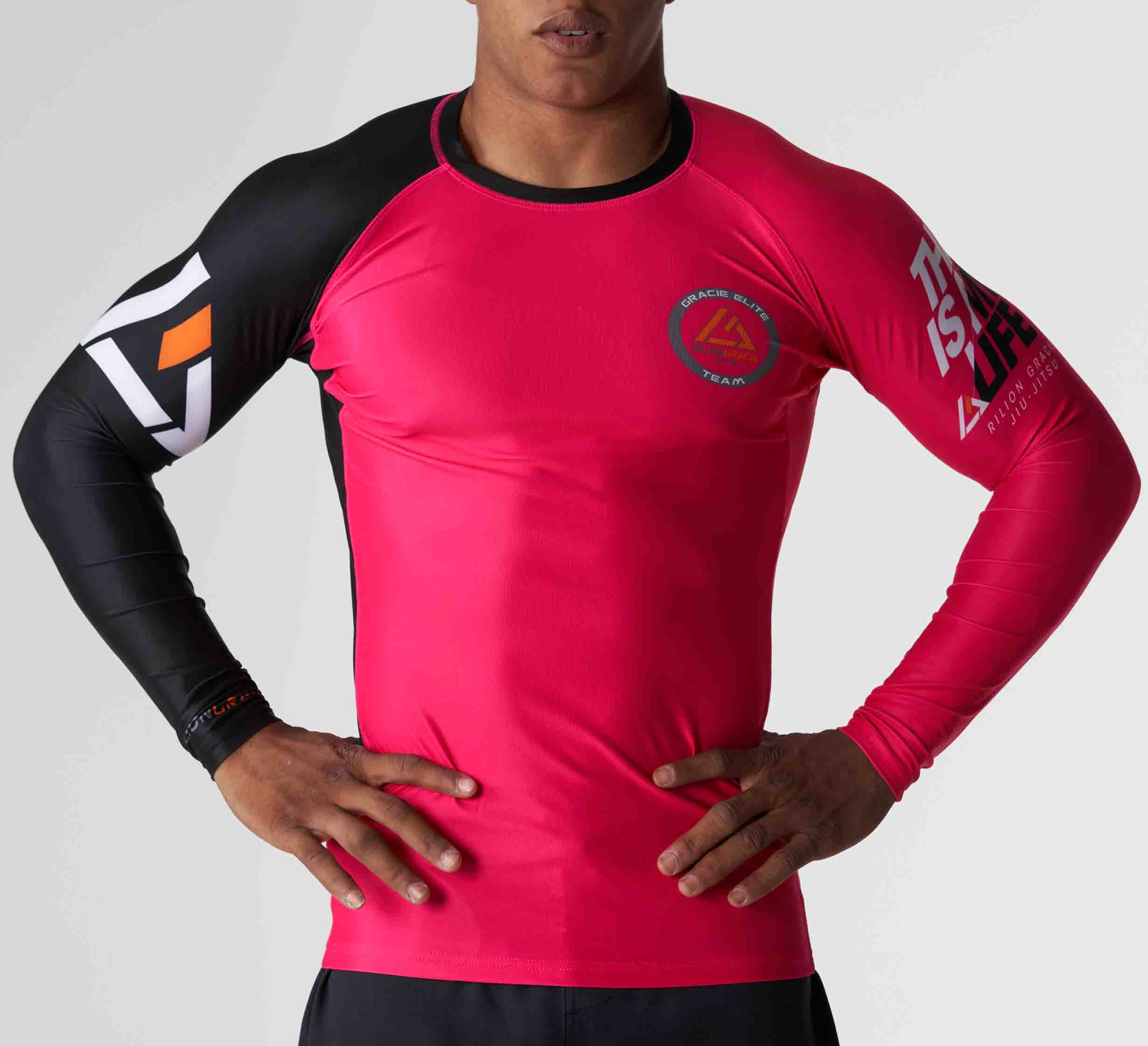 A woman wearing the Rilion Gracie Womens Rashguard Pink, a fitted long-sleeve top with pink body and black sleeves featuring logos, stands hands on hips against a plain background.