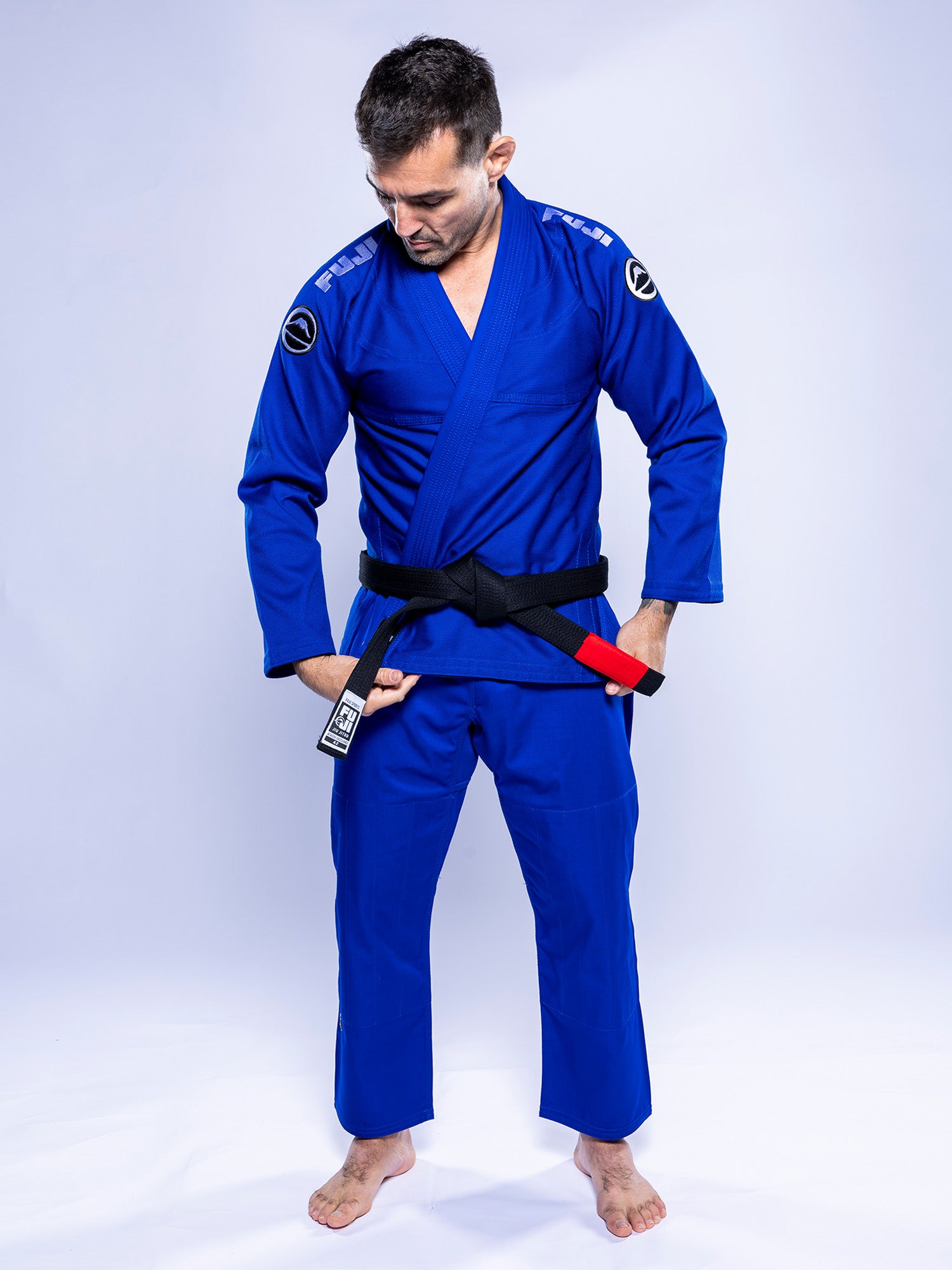 A man in the Baseline 2.0 BJJ Gi Blue White and a black belt stands barefoot on a white background, looking down and pointing at the tip of his belt.
