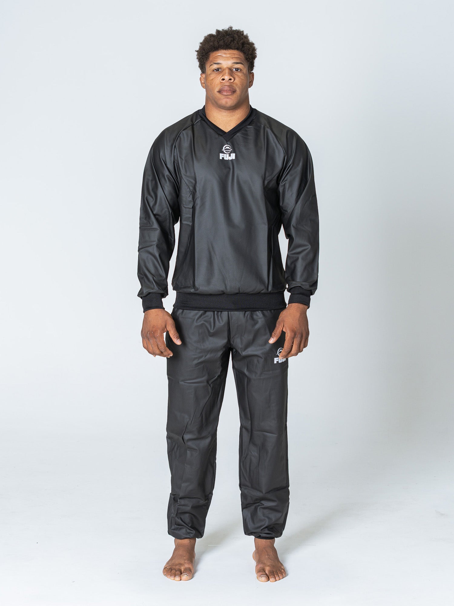 A person stands barefoot against a plain light background, wearing the Grappling Sauna Suit Black with visible Fiji logos on the jacket and pants. Their arms are relaxed by their sides.