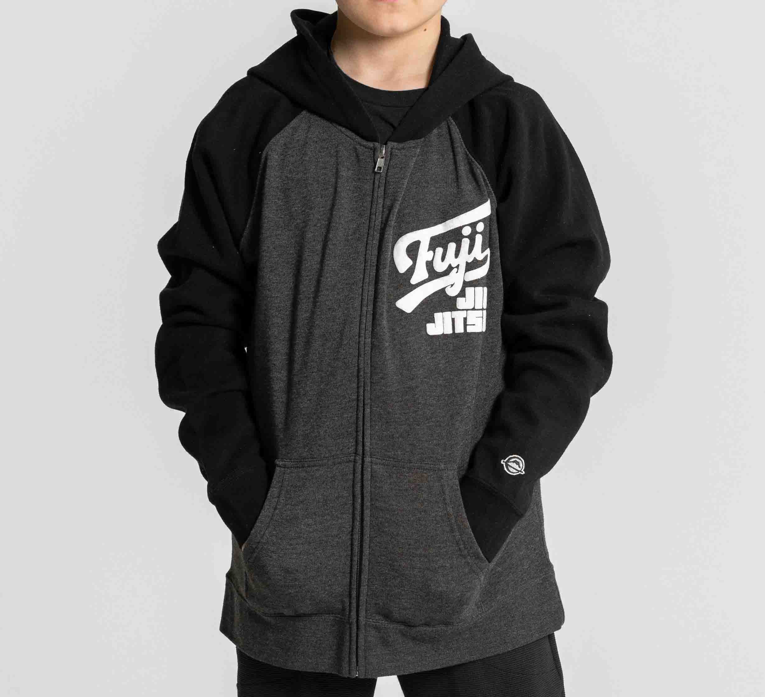 A child wears the Kids Jiu Jitsu Varsity Zip Hoodie in charcoal with “Fuji Jiu Jitsu” on the chest, hands in pockets, standing against a plain light background.