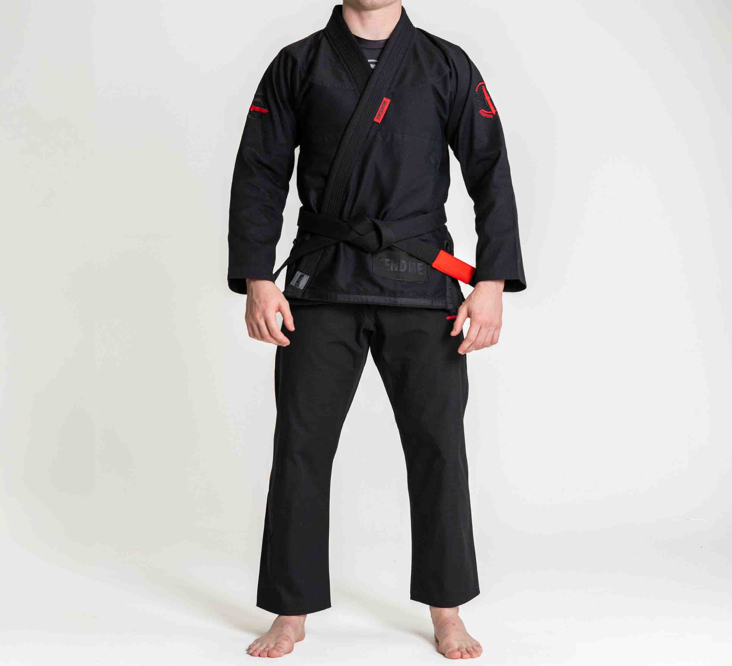 A person wearing the Suparaito BJJ Gi Sheepdog Response Edition Black with a black belt stands barefoot on a white background. Their face is not visible.