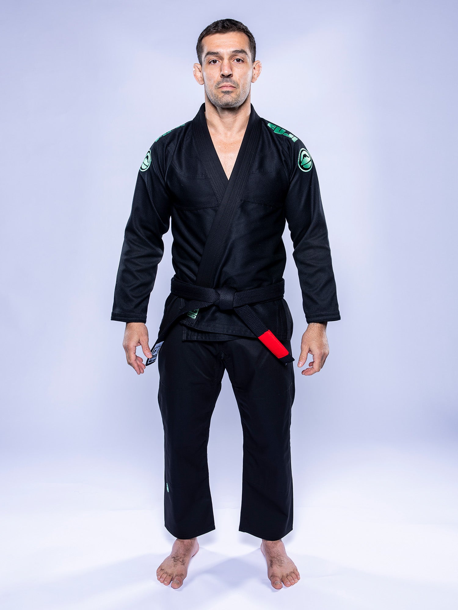 A person stands barefoot on a white surface, wearing the Baseline 2.0 BJJ Gi Black Mint and a red-and-black belt. The background is plain light purple, and they face forward with a neutral expression.