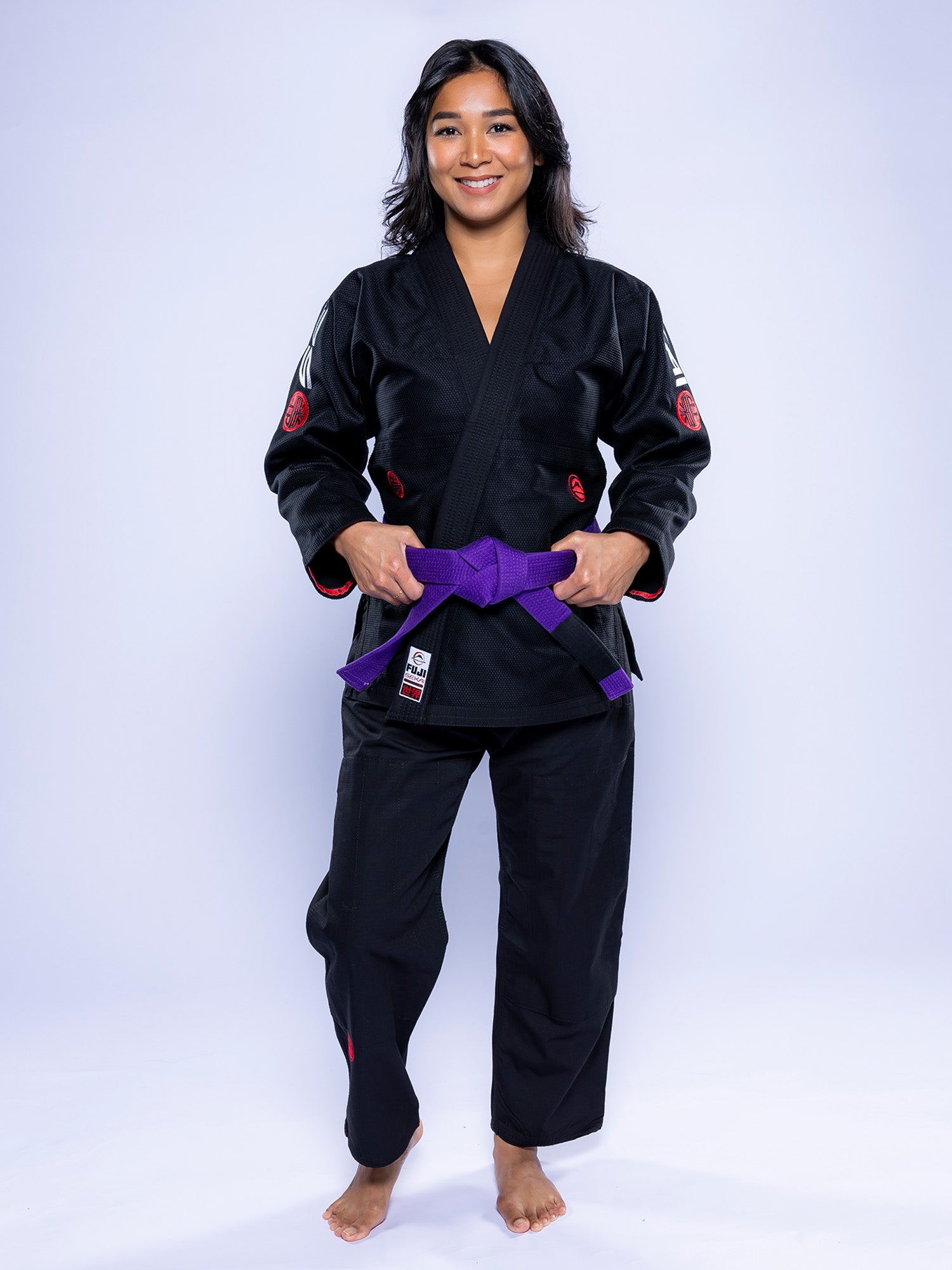 A woman stands barefoot on a white background, smiling and wearing the Womens SEKAI 3.0 BJJ Gi Black Grey with a purple belt tied around her waist.