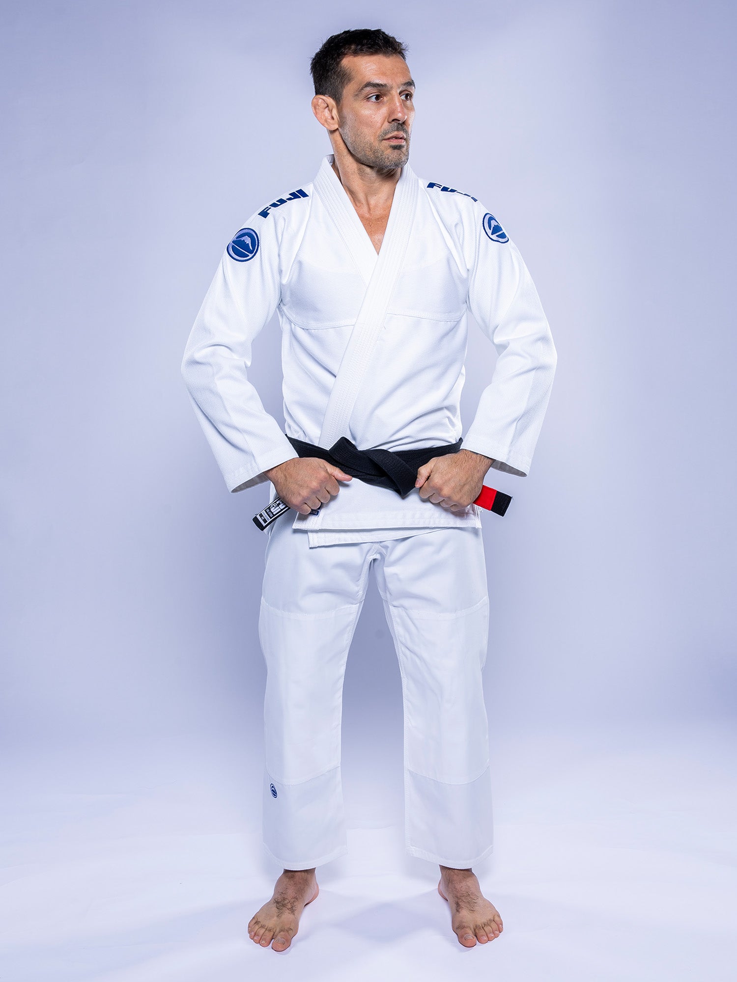 A man wearing the Baseline 2.0 BJJ Gi White Navy stands barefoot on a white background, holding his black belt with both hands and gazing slightly to the side.