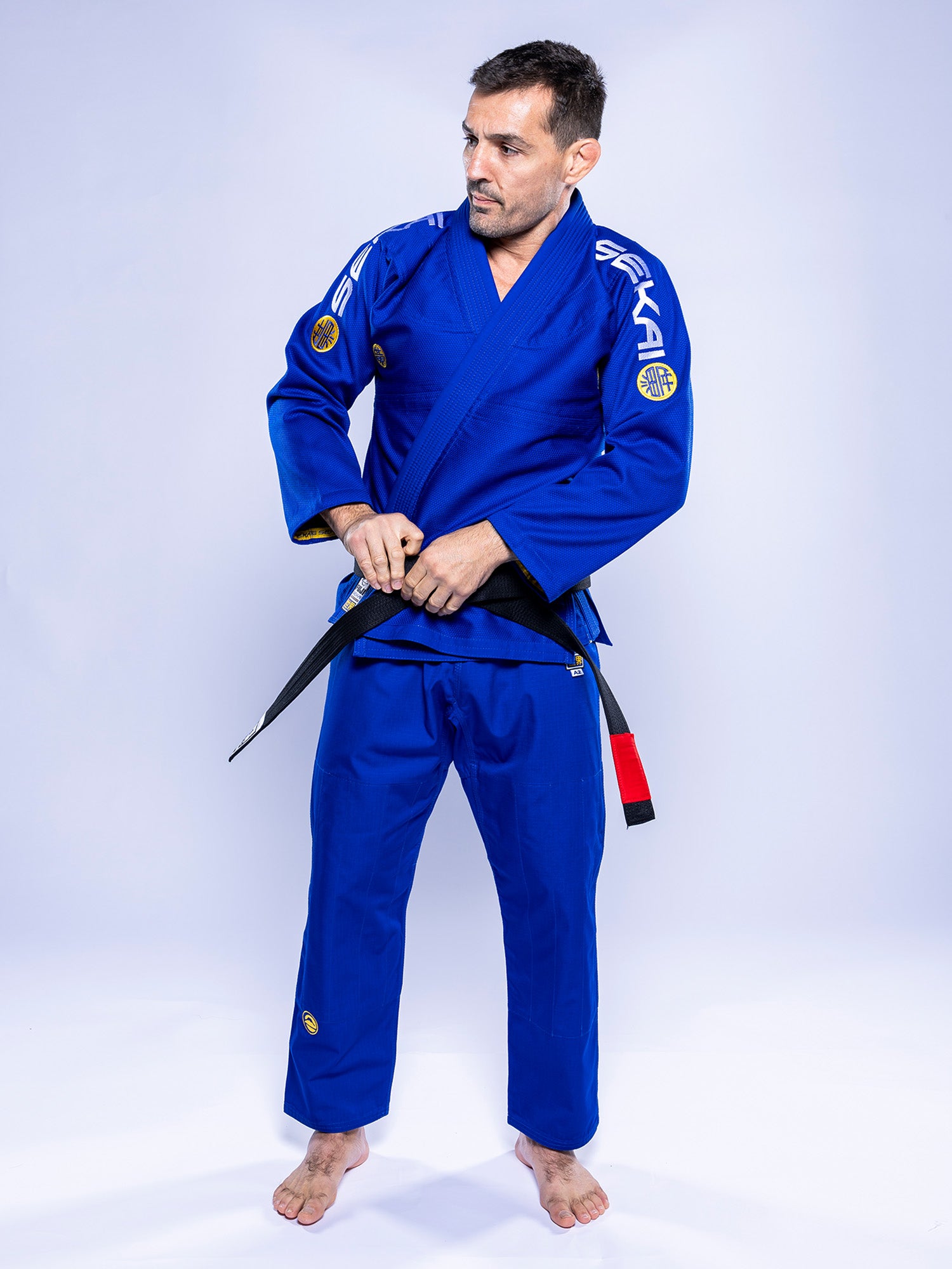 A man in the SEKAI 3.0 BJJ Gi Blue Yellow stands barefoot on a white background, looking to the side as he ties a black belt around his waist.