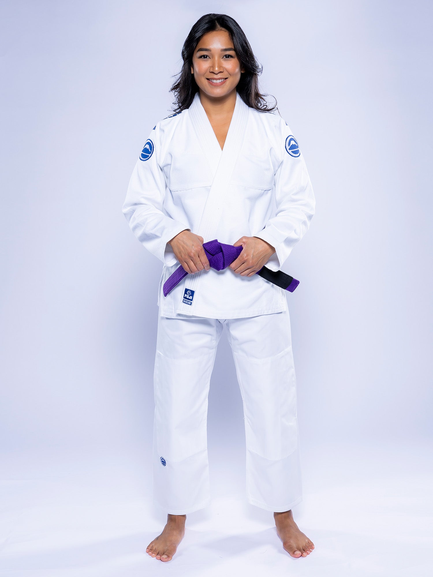 A woman stands barefoot on a white surface, smiling and wearing the Womens Baseline 2.0 BJJ Gi White Navy with patches on the sleeves, holding a purple belt in both hands.