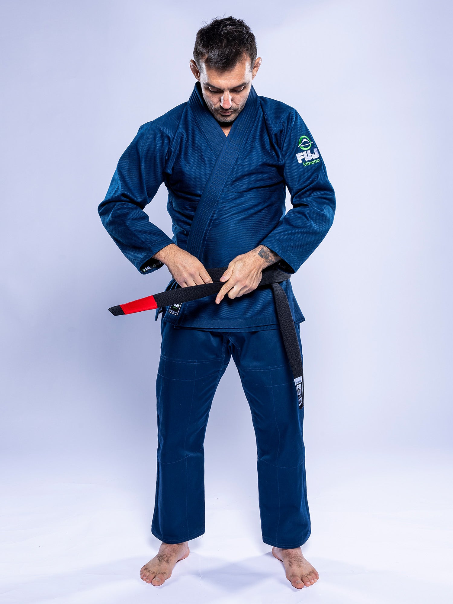 A man stands barefoot on a white background, looking down as he ties a black belt with a red bar around his waist while wearing the Lightweight BJJ Gi Navy.