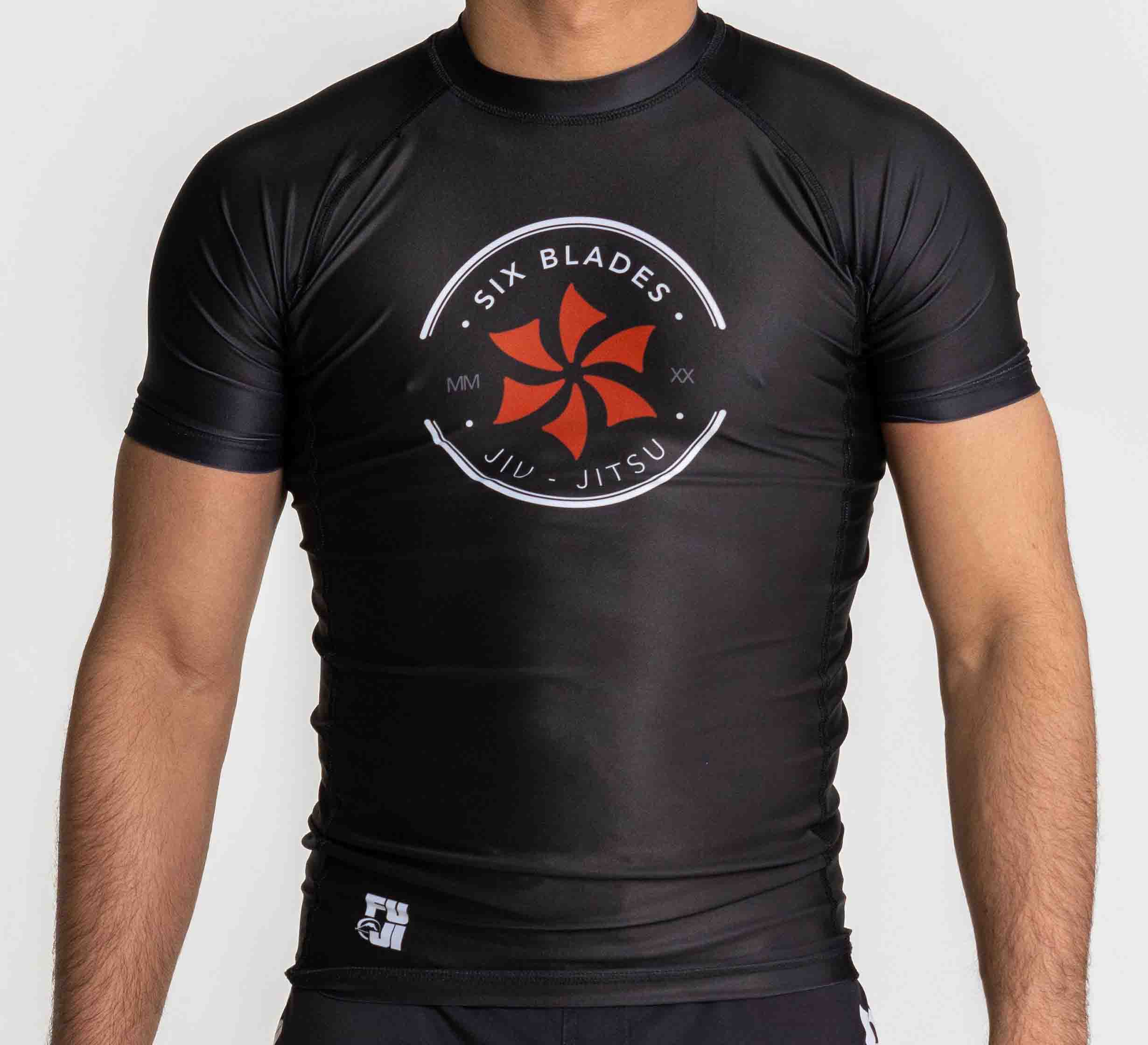 A person wearing the Six Blades Short Sleeve Rashguard Black, featuring “Six Blades Jiu Jitsu” and a red six-blade logo on the front, short sleeves, and white branding near the waist. The person’s head is not visible.