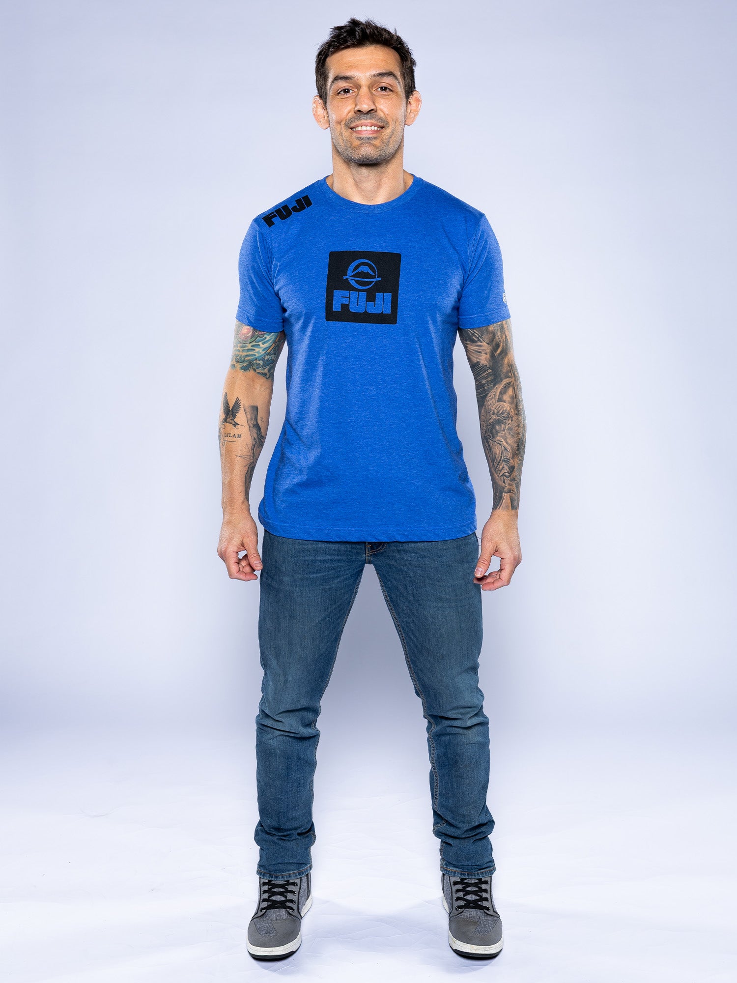 A man with tattoos on both arms smiles, wearing the Elevate Your Journey Shirt Blue, blue jeans, and gray sneakers against a plain light background.