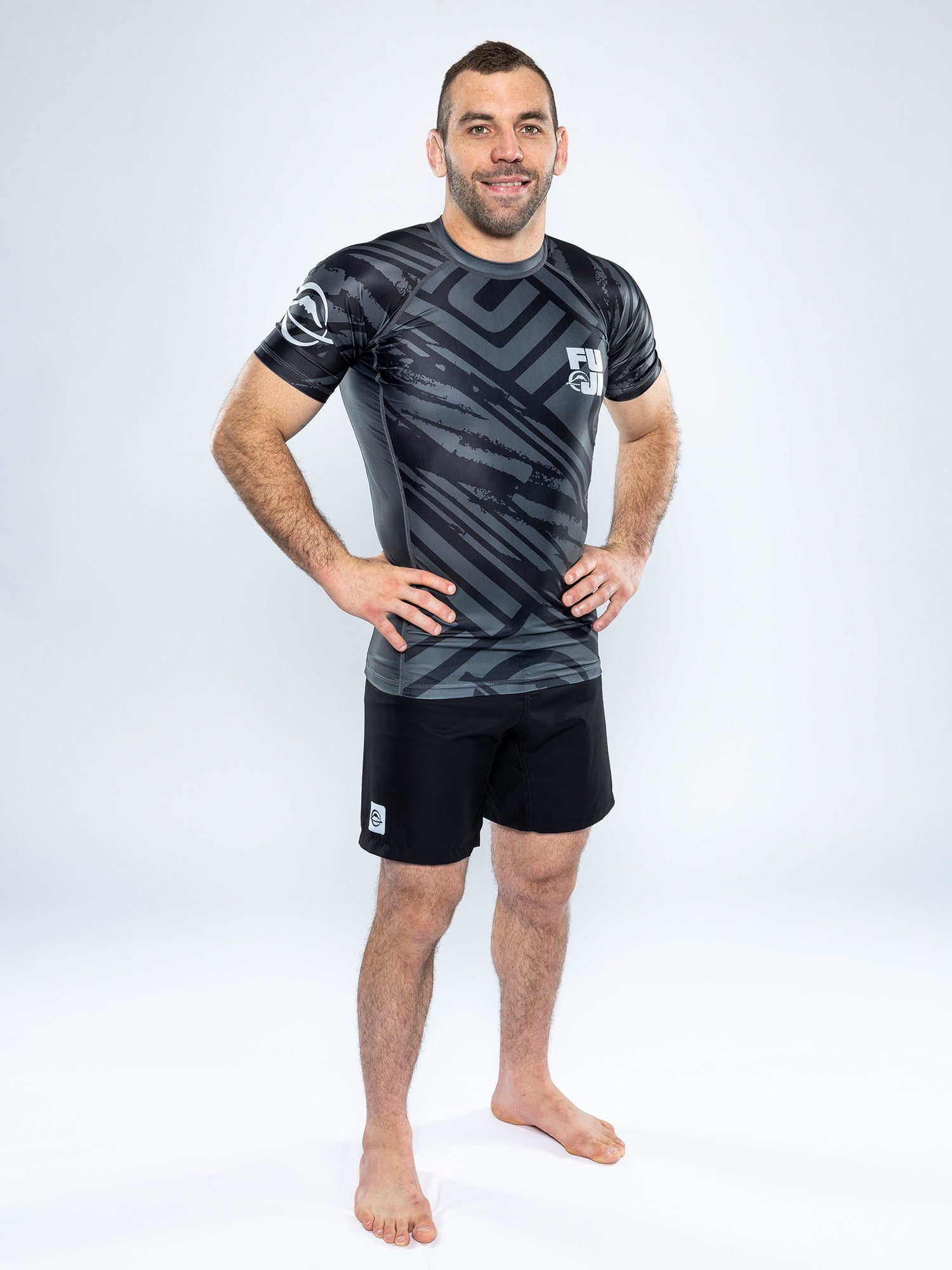 A man stands barefoot on a white background, smiling with his hands on his hips, wearing the Battle Flex Lite Rashguard Black and black shorts featuring a geometric pattern.